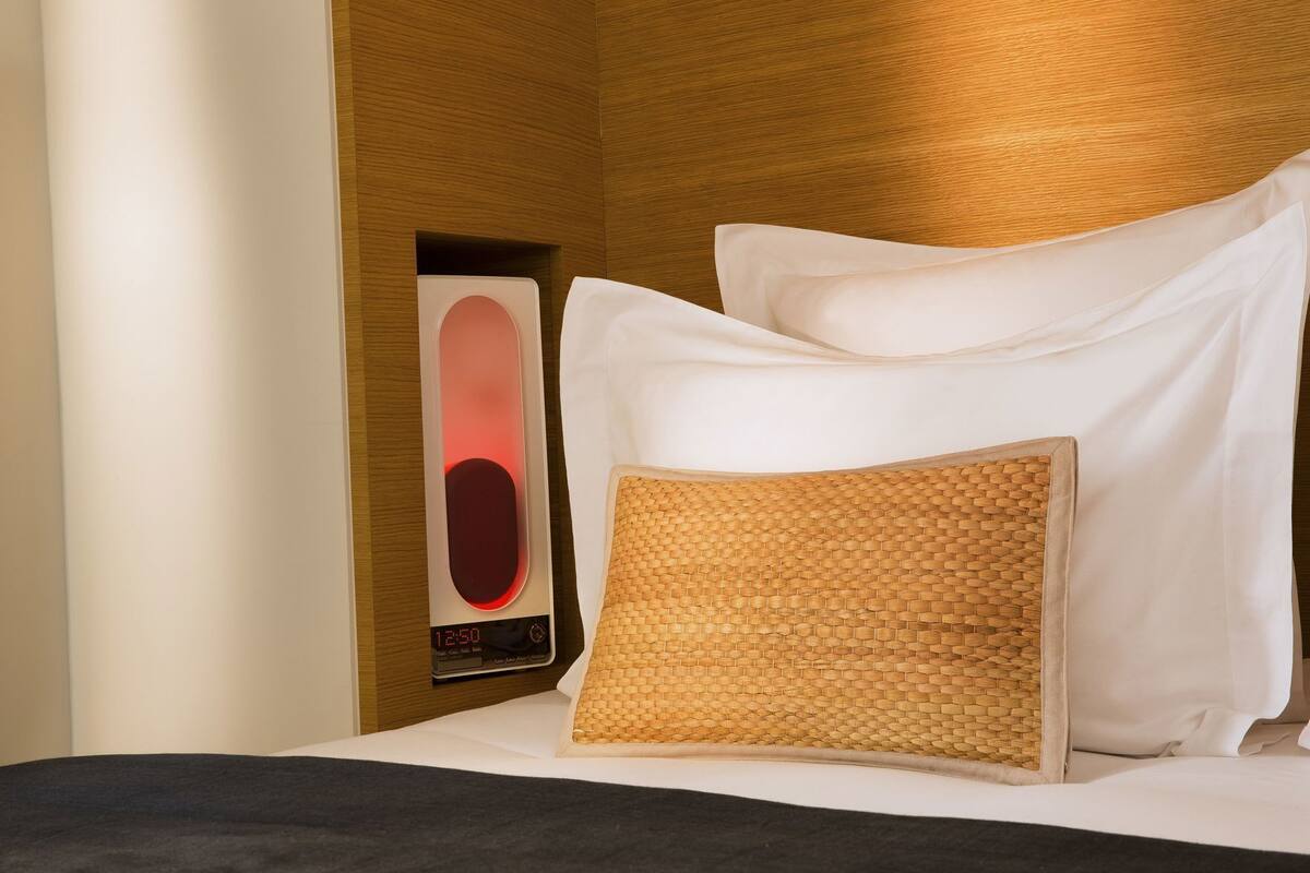 Una cama bien hecha con sábanas blancas, un cojín tejido y un reloj digital sobre una mesita de noche de madera.
