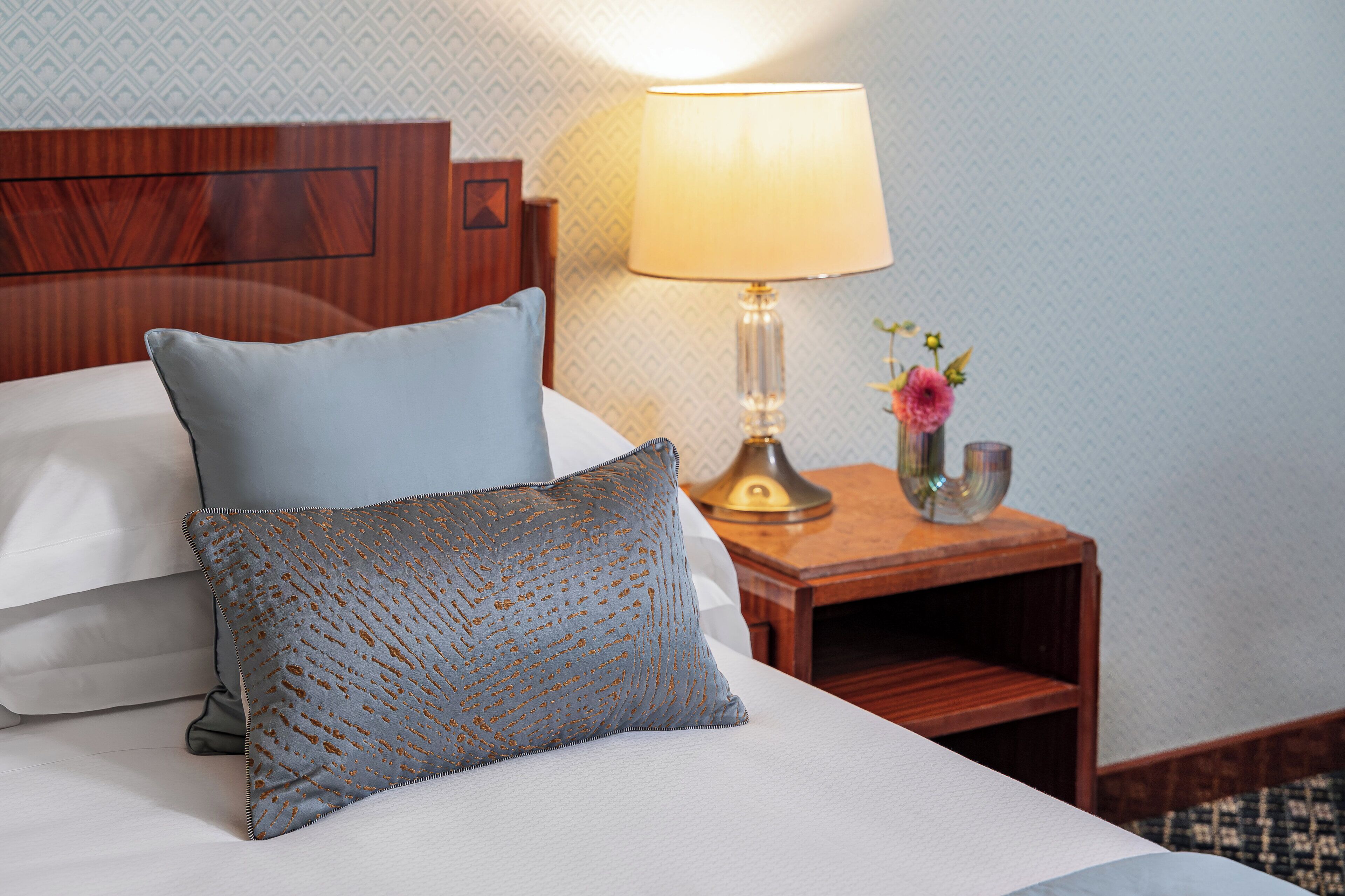 A neatly made bed with two pillows, a bedside lamp, and a small vase with flowers.