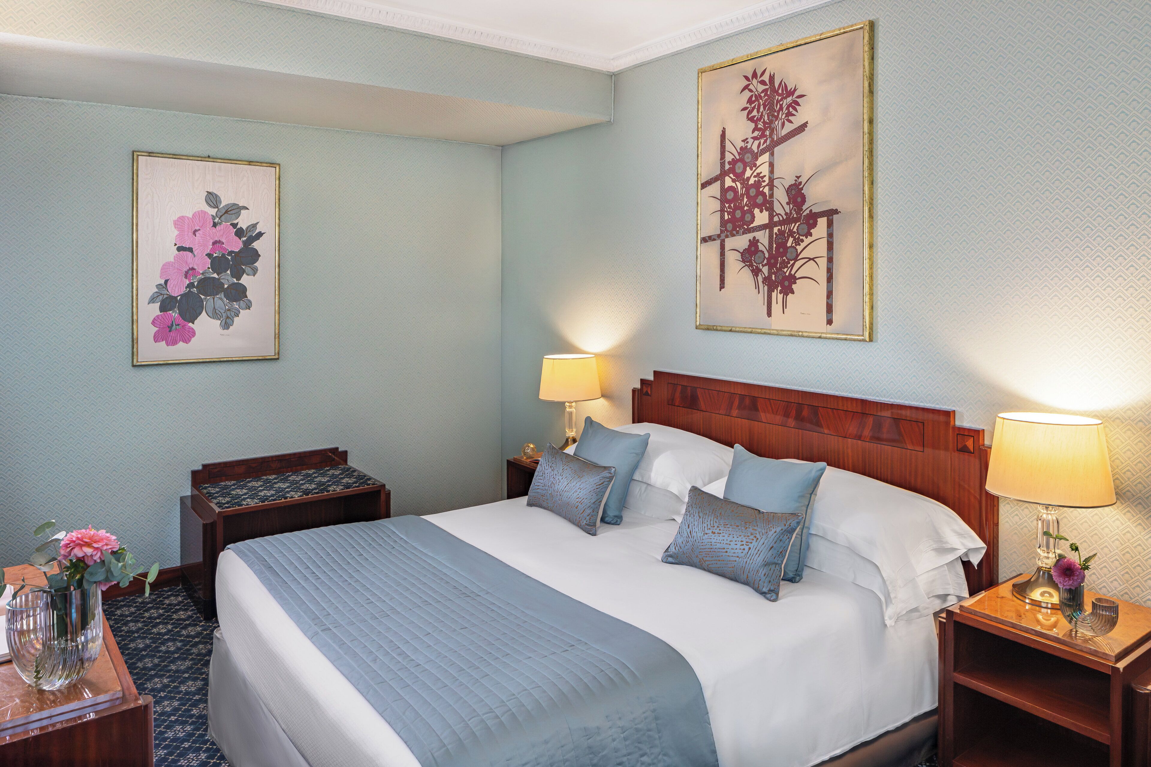 Una camera d'albergo ordinata con un letto grande, due comodini con lampade e due quadri floreali appesi alle pareti.