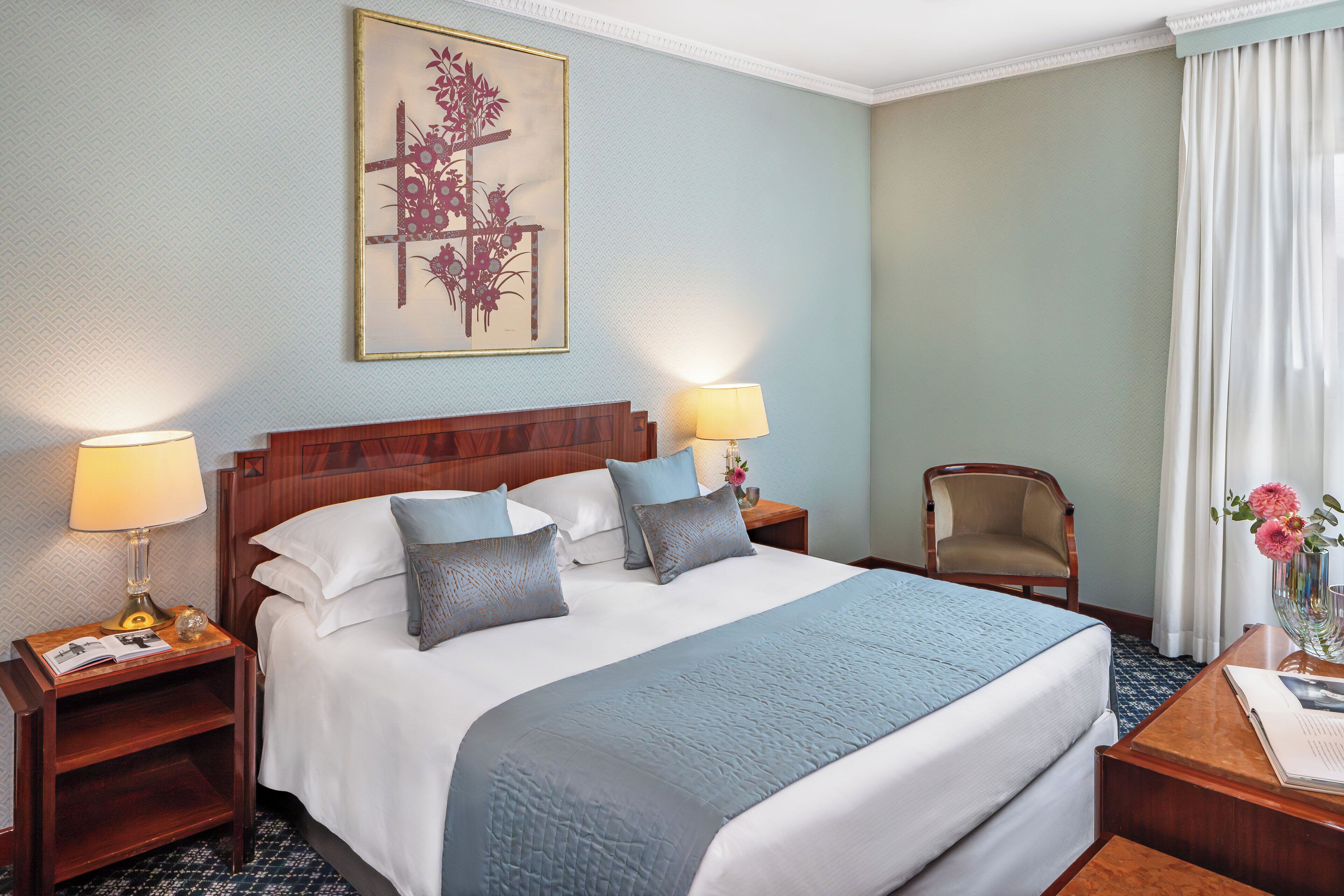 Uma cama bem arrumada com um cobertor azul, duas luminárias de cabeceira, uma mesinha de cabeceira de madeira, uma cadeira e uma obra de arte emoldurada na parede.