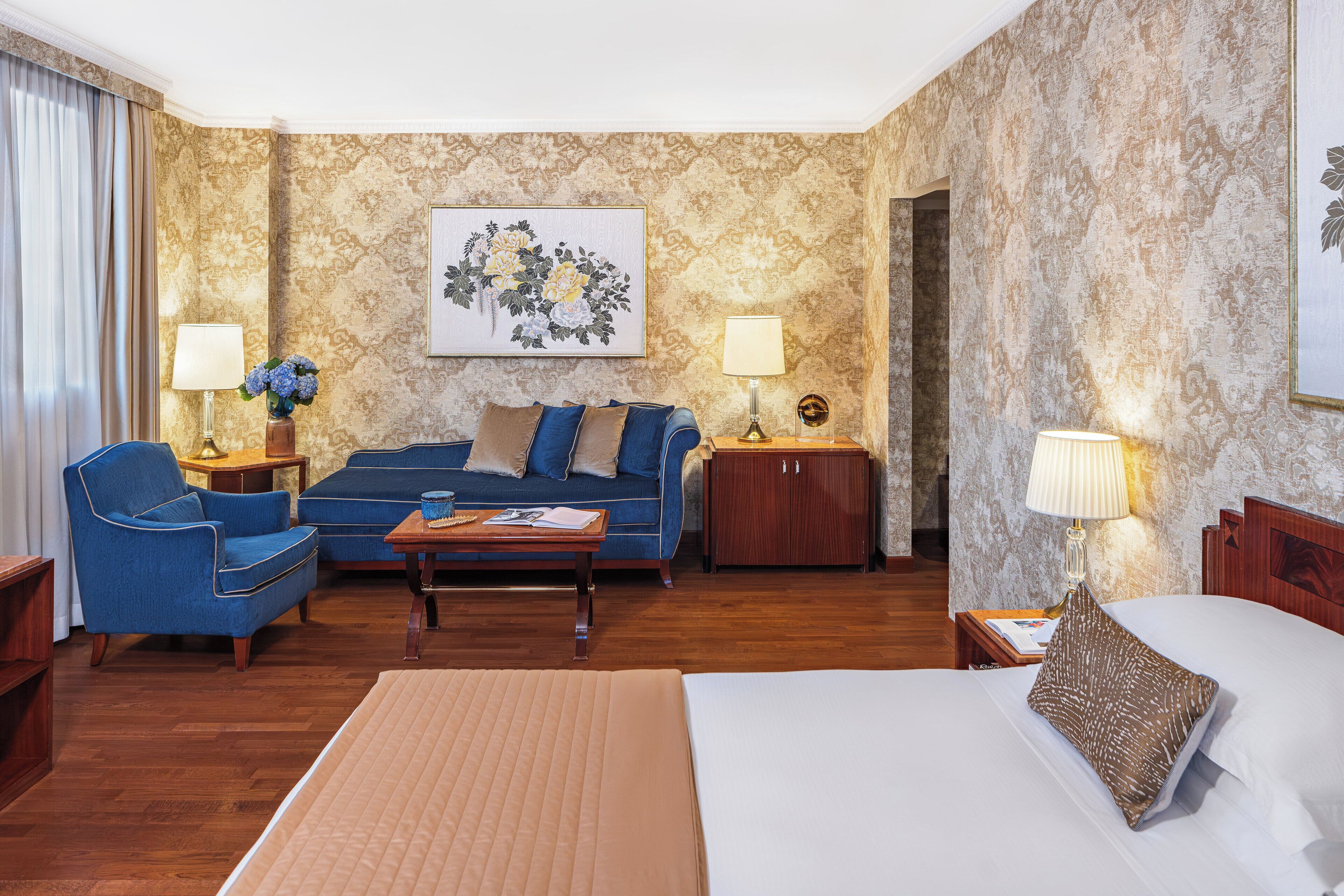 Habitación de hotel con cama, sofá, sillón, una mesita y dos mesitas de noche con lámparas.