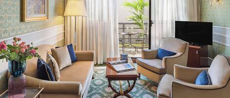 A hotel room with a sofa, armchairs, a coffee table, a TV, a lamp, and a window with curtains.