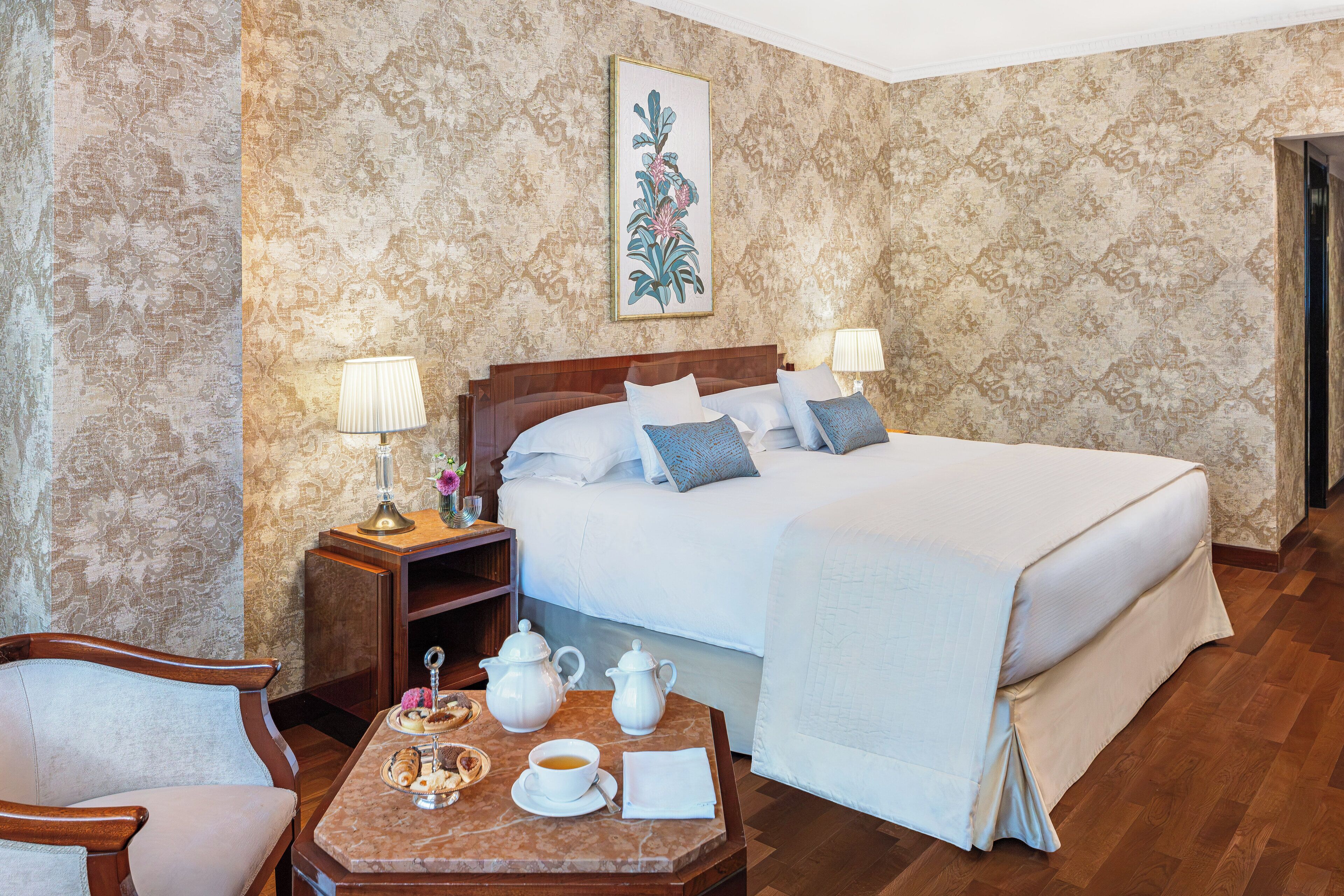 A hotel room with a large bed, two bedside lamps, a wooden nightstand with a lamp, a small table with a teapot, cups, and pastries.