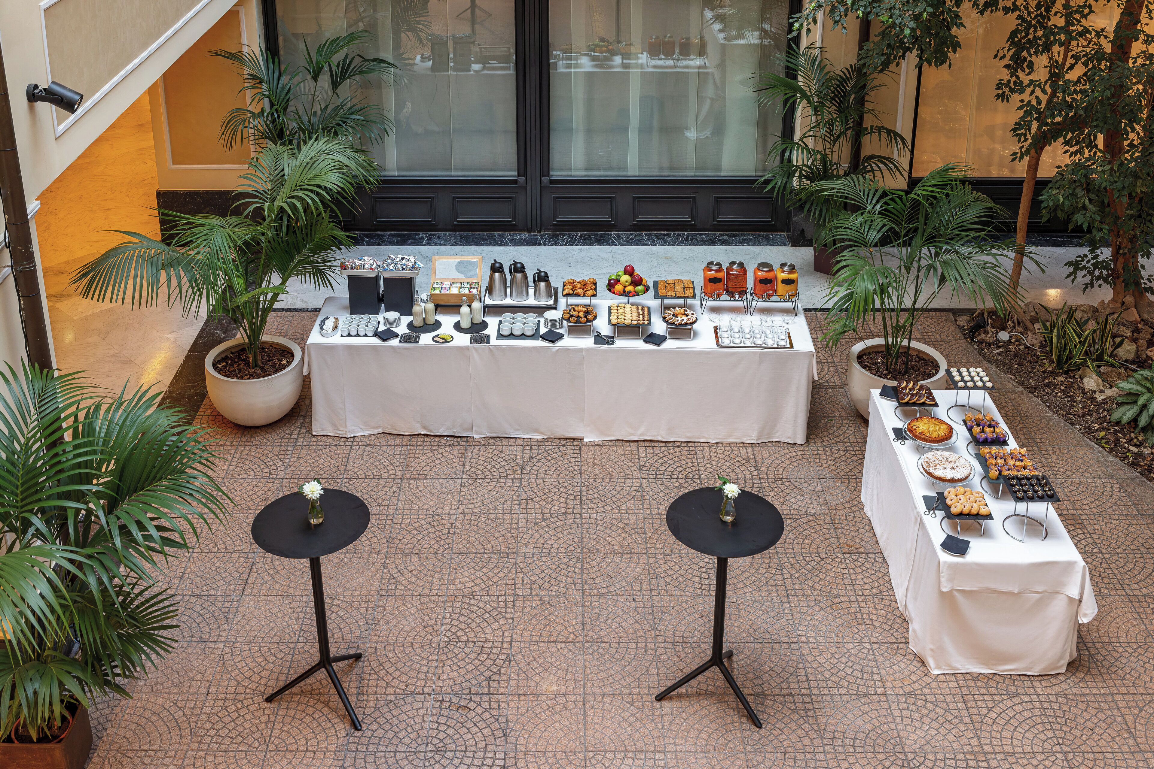 An outdoor buffet setup with a variety of food items and beverages, surrounded by potted plants and a tiled floor.