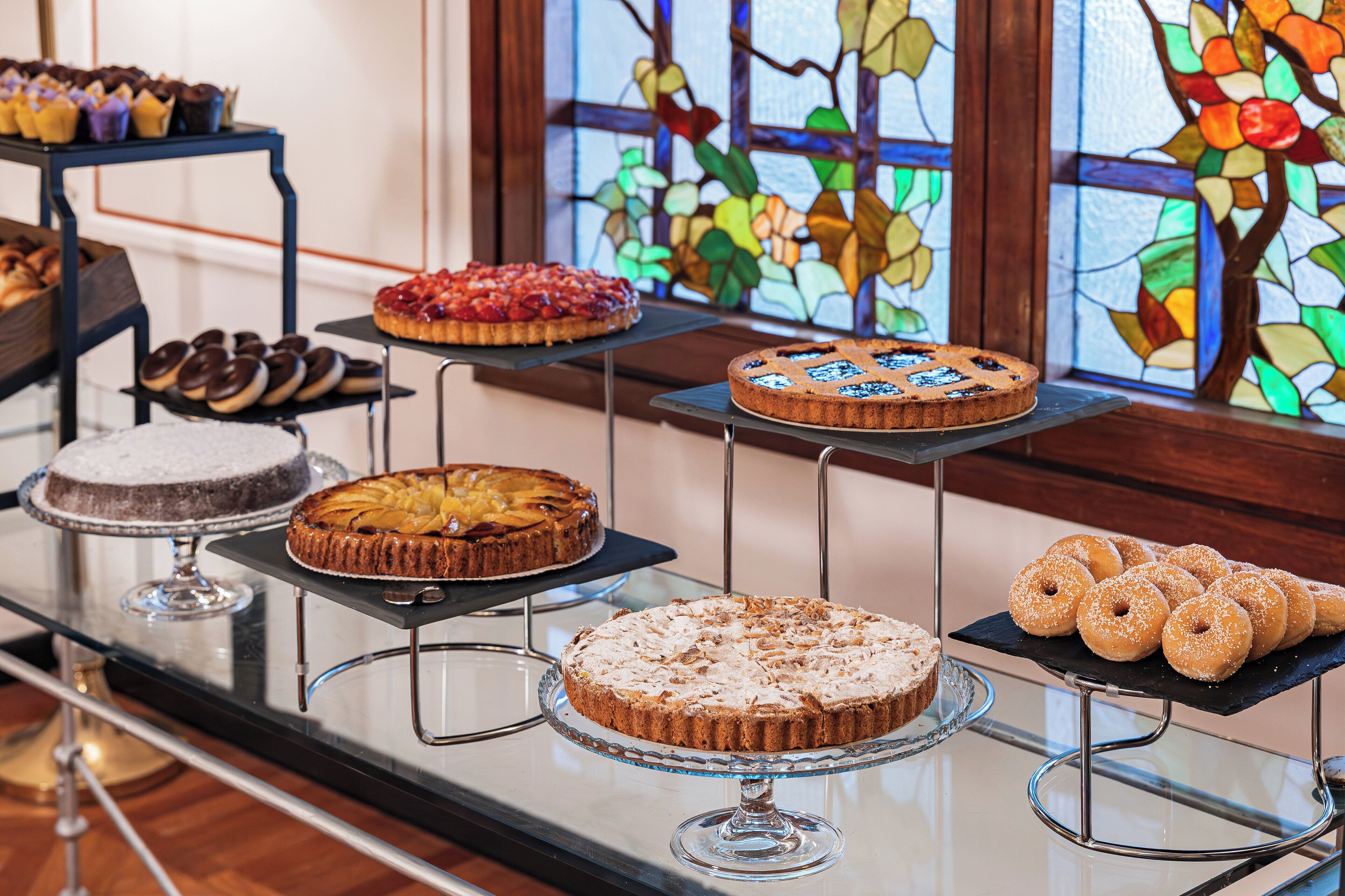 A display of various cakes and pastries on stands in a room with a stained glass window in the background.
