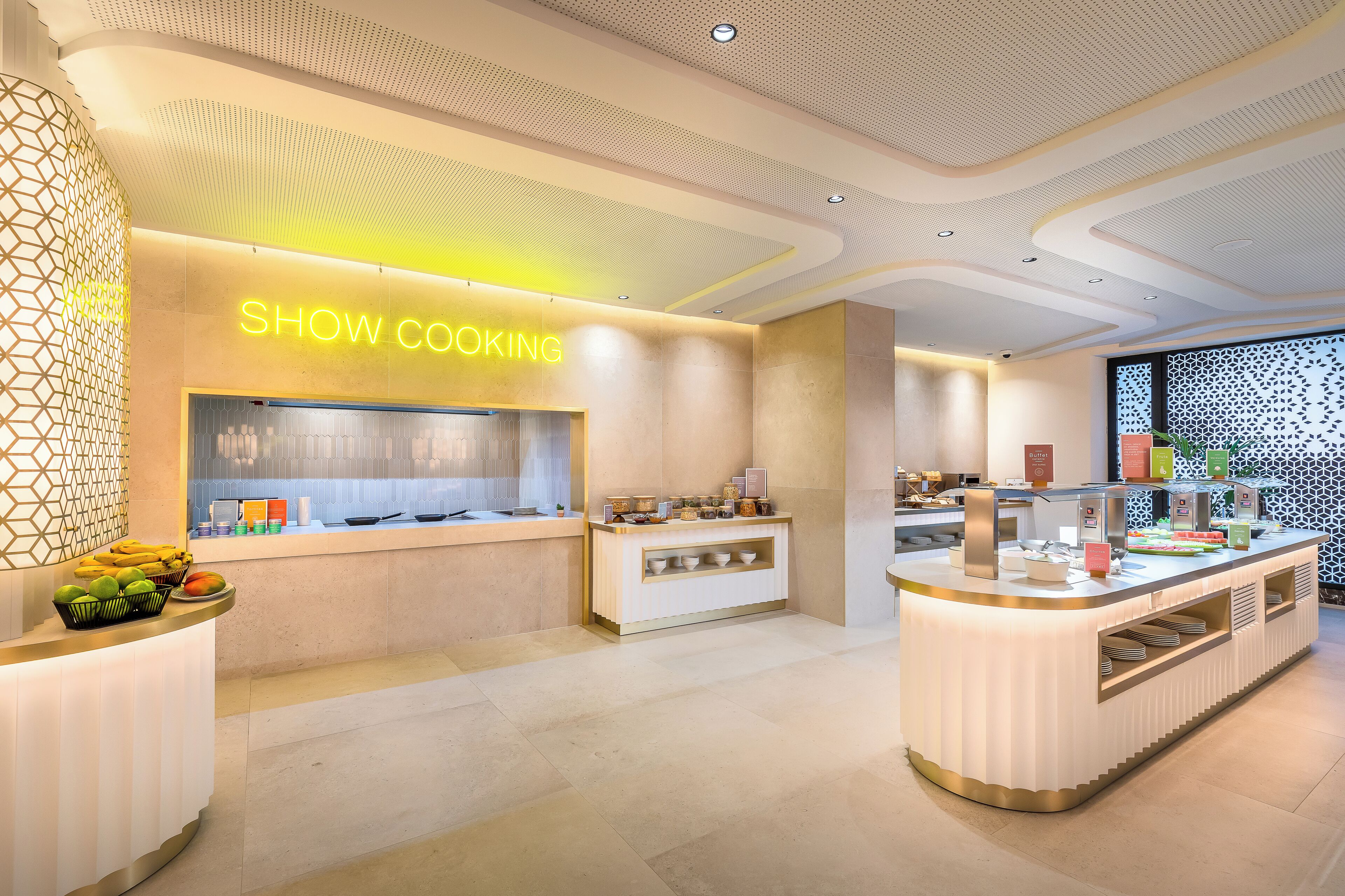 Un hall d'hôtel moderne avec une enseigne indiquant un espace de « CUISINE DÉMONSTRATIVE », un présentoir de fruits et un comptoir proposant divers produits alimentaires.