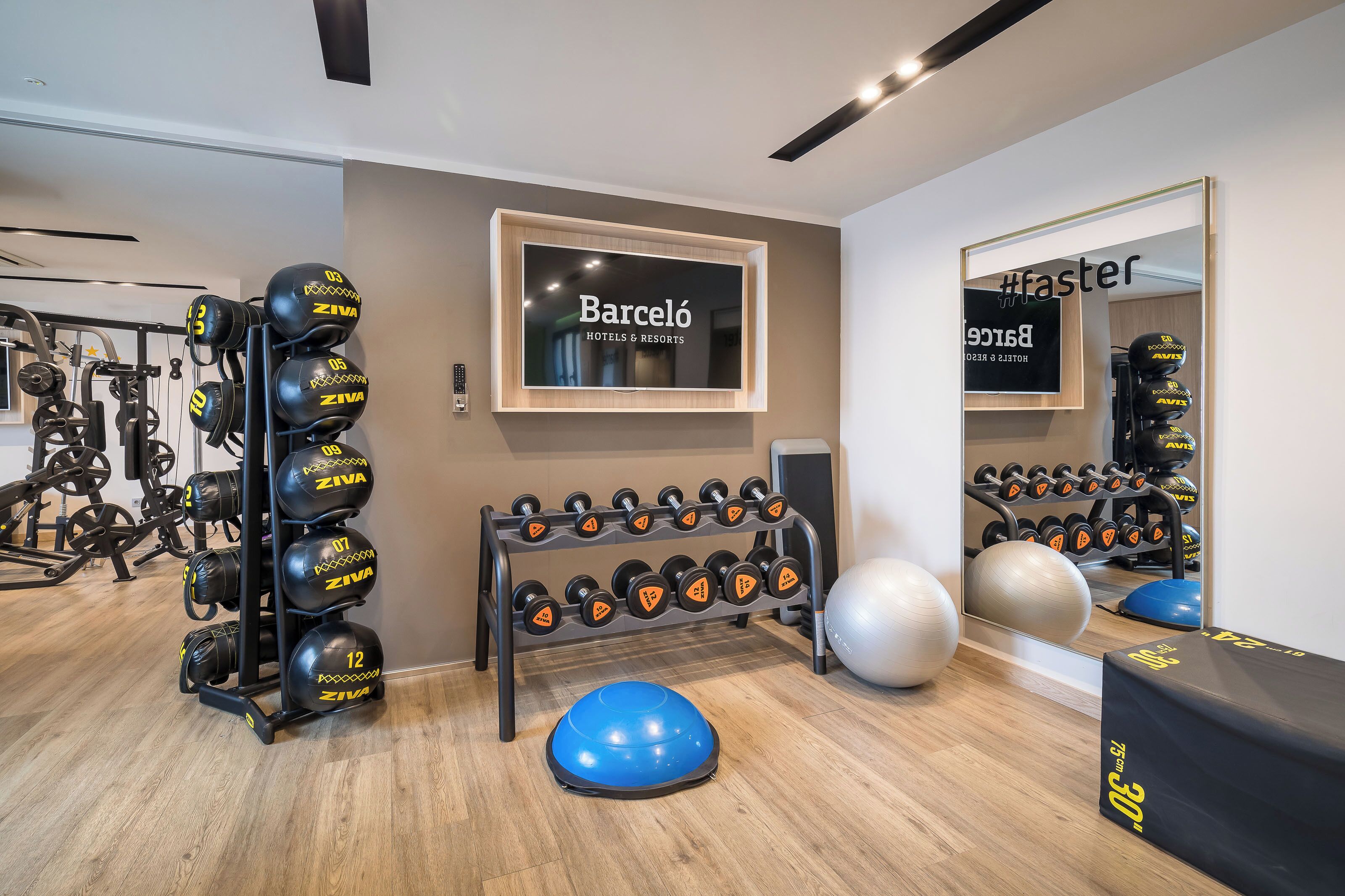 Ein Fitnessraum mit Trainingsgeräten, einem Spiegel und einem an der Wand montierten Fernseher, der den Namen des Hotels zeigt.
