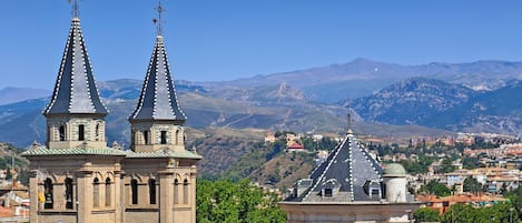 A historic building with two prominent towers and a dome, surrounded by a mountainous landscape and a town below.
