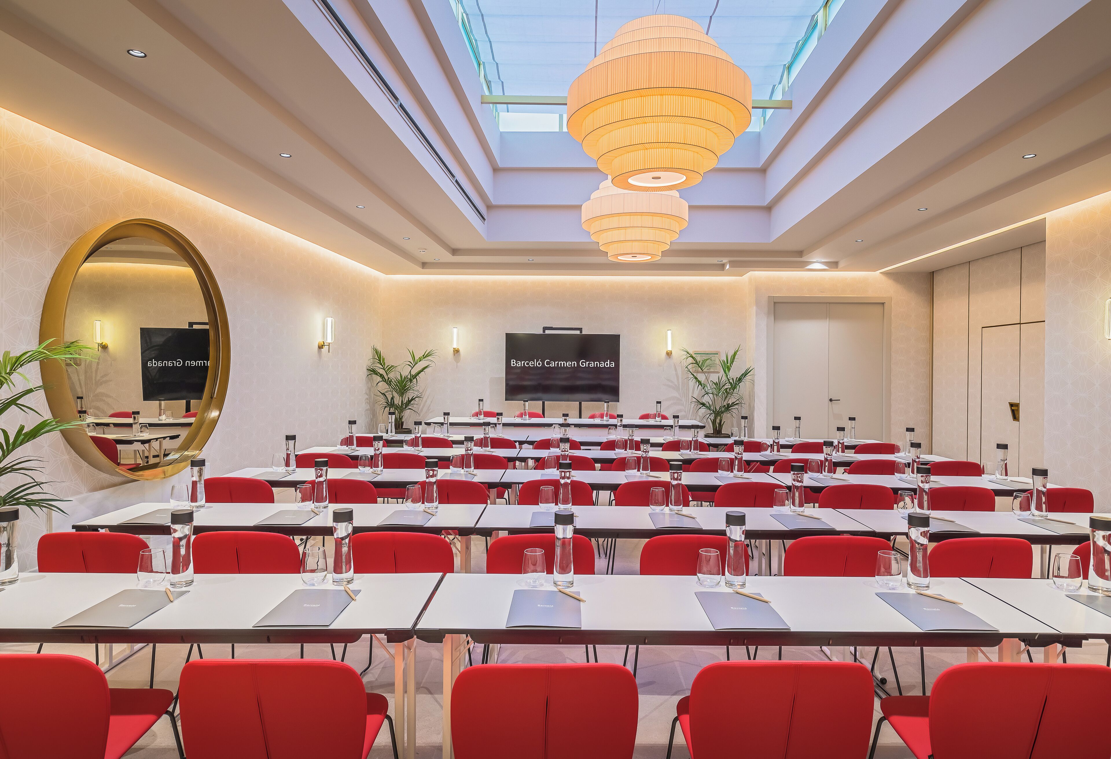 A conference room with multiple tables and chairs, a large screen, and a ceiling with a skylight and pendant lights.