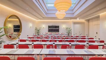 A conference room with multiple tables and chairs, a large screen, and a ceiling with a skylight and pendant lights.