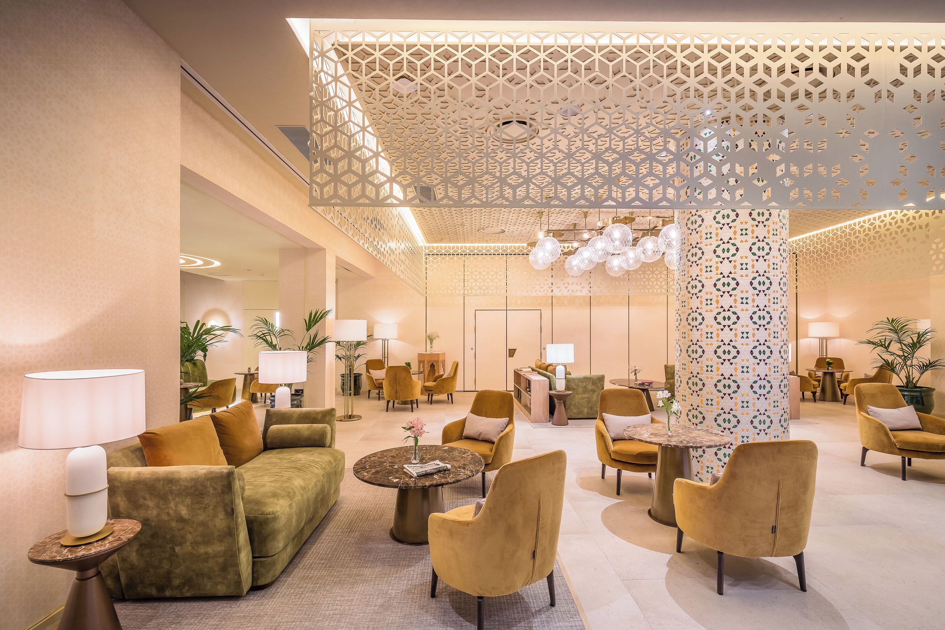 Un amplio vestíbulo con techo alto, diseño de techo estampado y una columna central con motivos decorativos. Hay varias zonas de estar con sofás y sillones, y una mesa de centro.