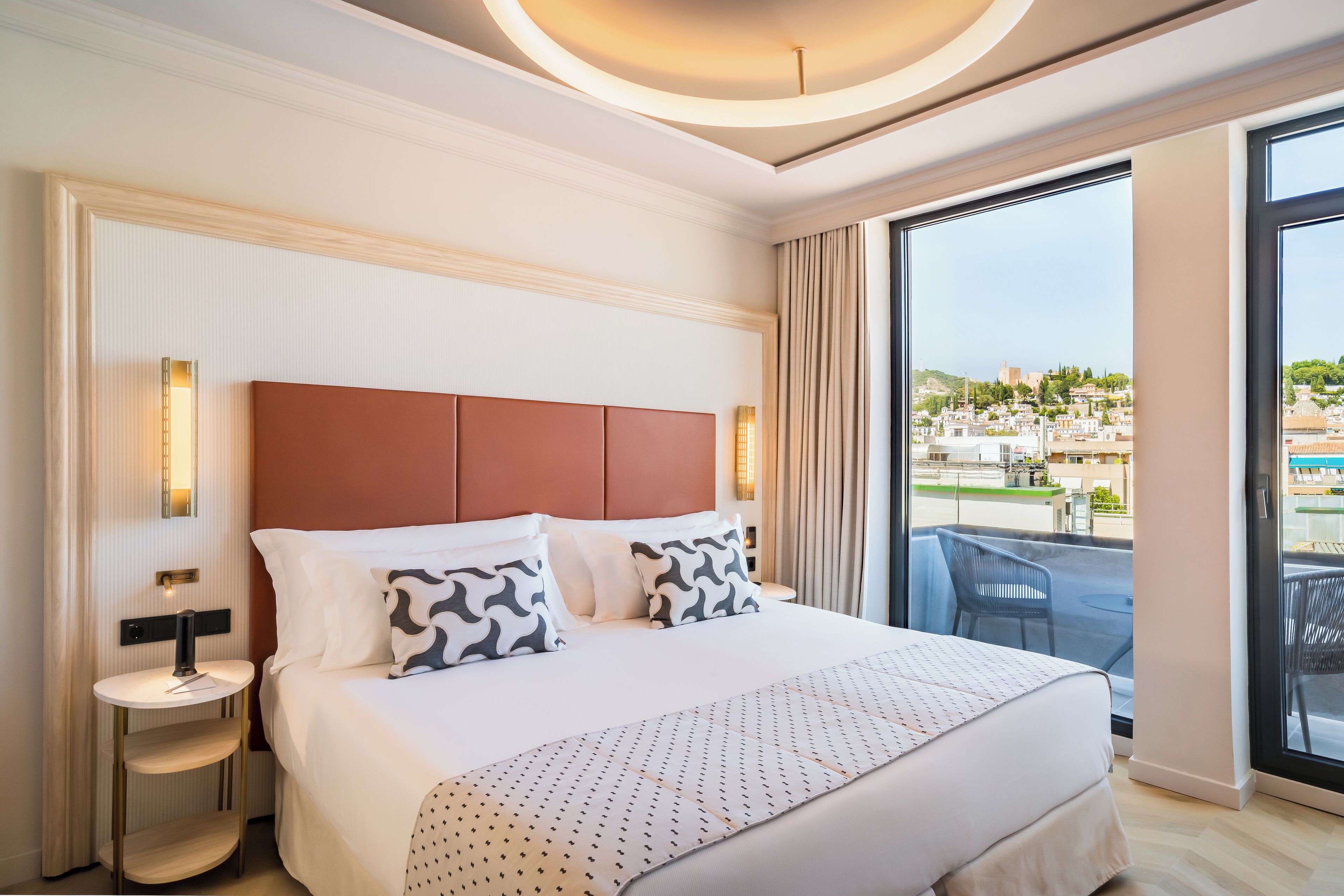 Une chambre d’hôtel avec un grand lit, une table ronde et une vue sur le paysage urbain, visible à travers de grandes fenêtres.