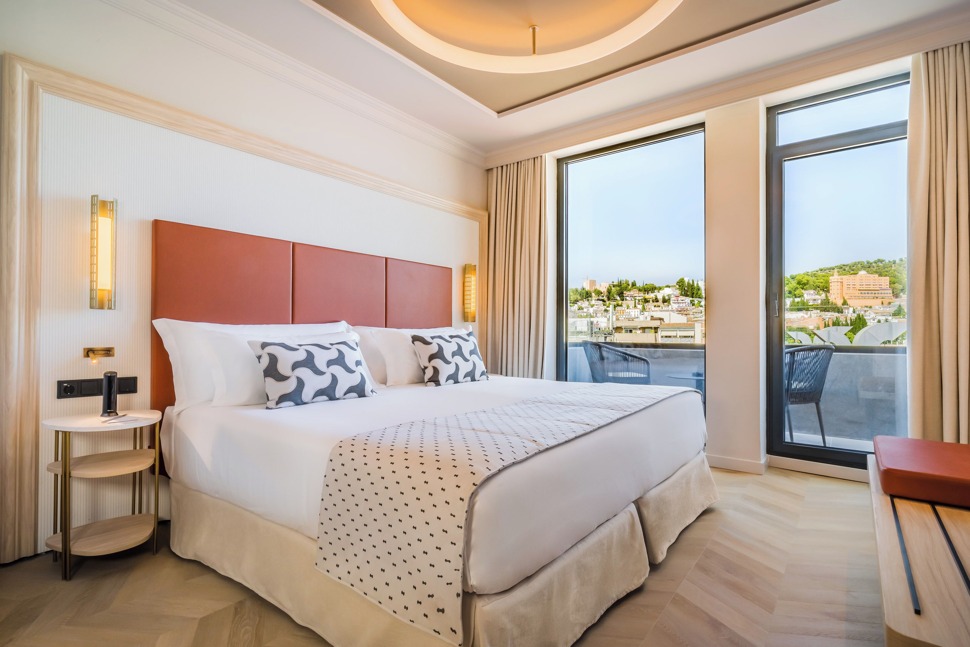 A hotel room with a large bed, a bedside table, a view of the city through large windows, and a balcony with outdoor seating.