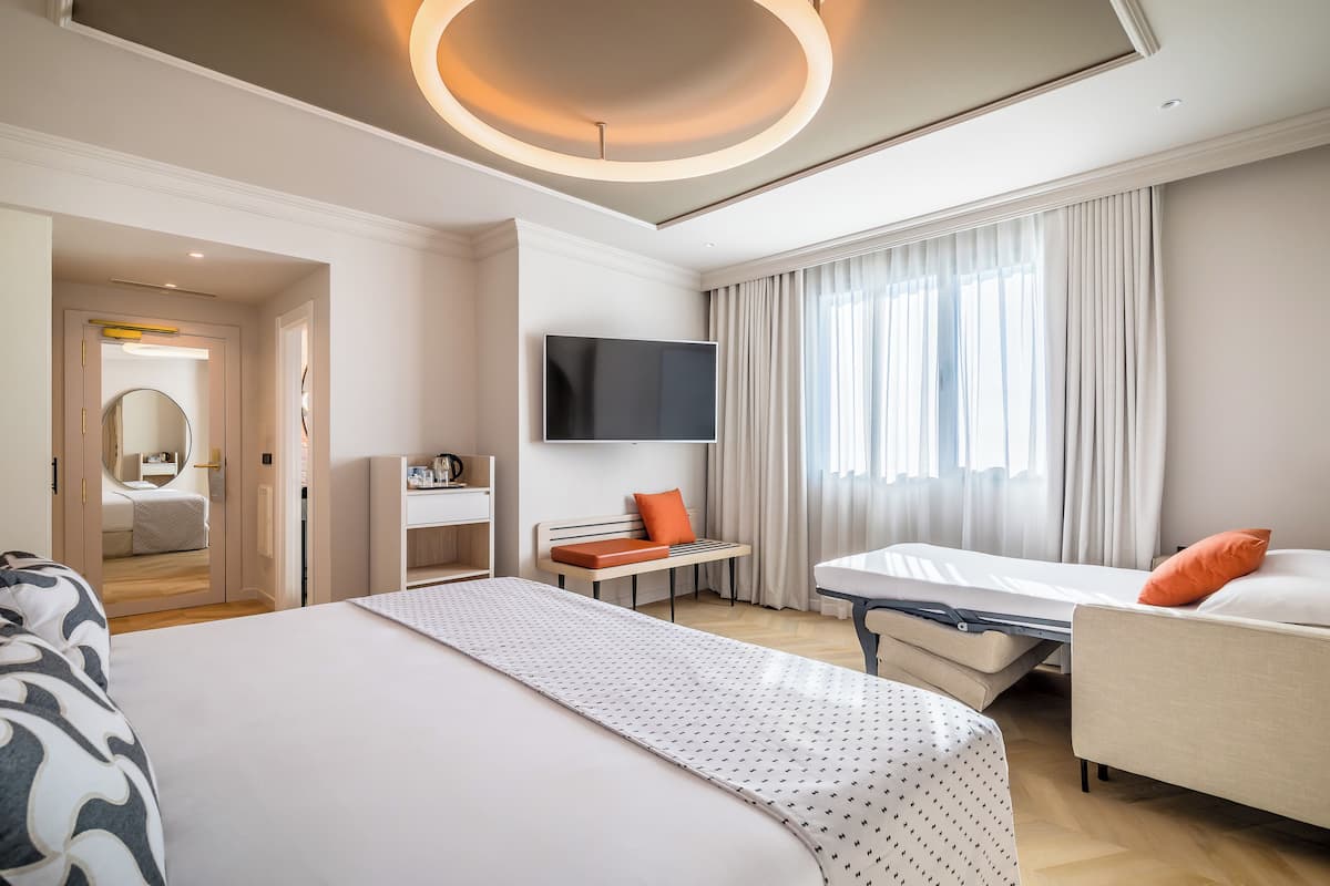 A hotel room with a large bed, a bedside table, a TV, a desk, and a window with curtains.
