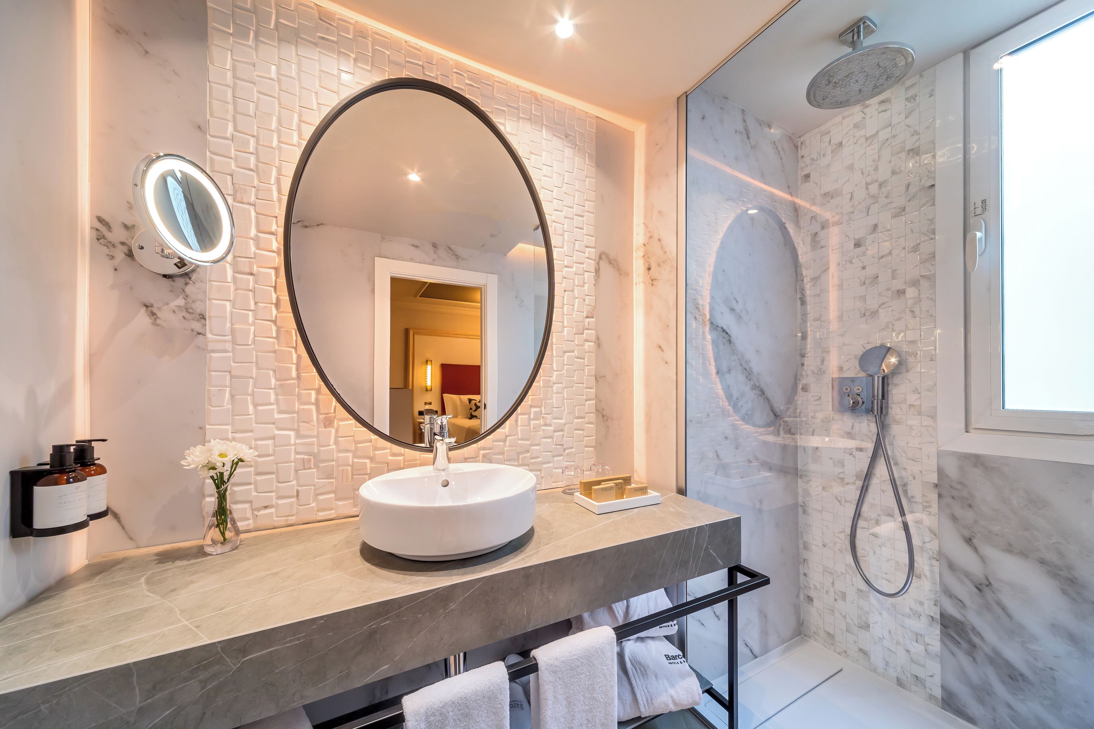 Un baño moderno con un espejo redondo, un lavamanos blanco y una ducha con mampara de vidrio.