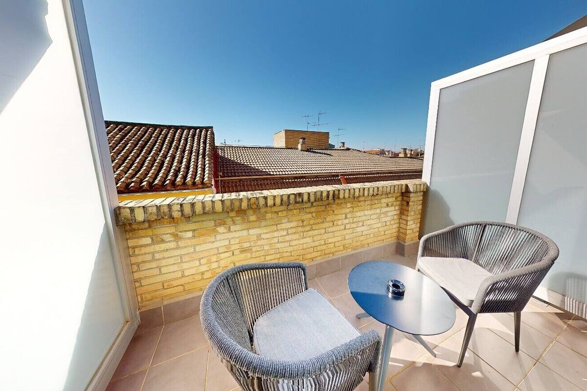 A small outdoor patio with two wicker chairs and a round table, a brick wall, and a tiled roof in the background.