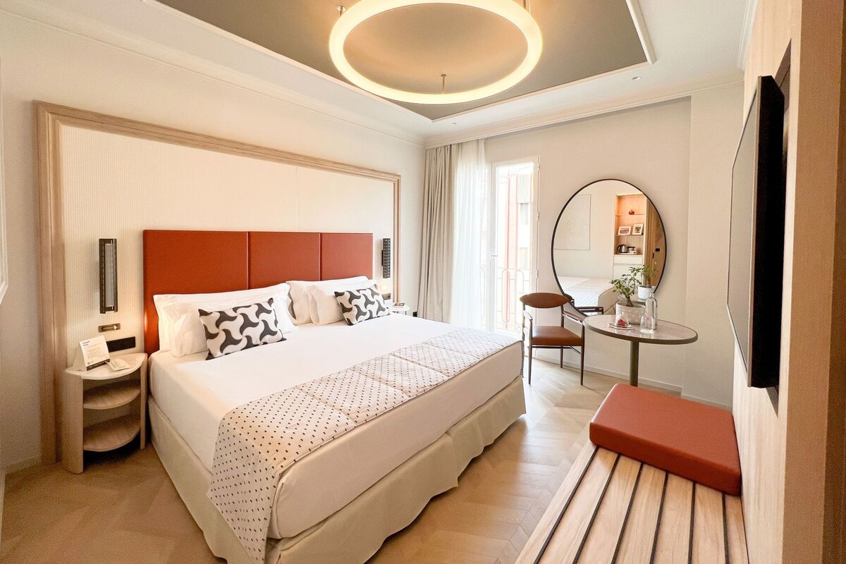 Habitación de hotel con una cama grande, mesitas de noche, un espejo redondo, una pequeña mesa con un florero y un banco rojo.