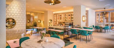A modern restaurant with round tables, green chairs, and a bar area with shelves of bottles.