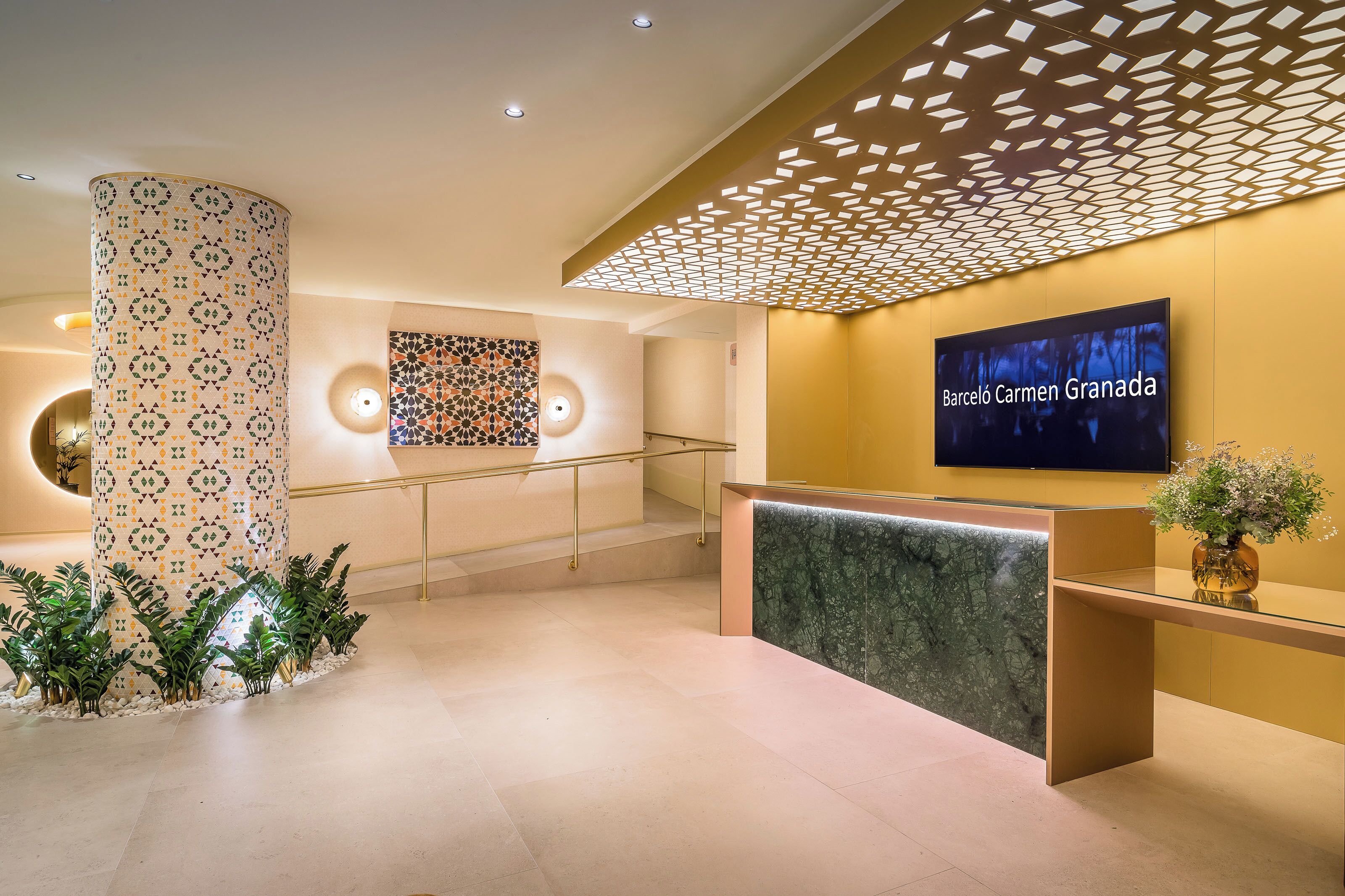 Una moderna zona de recepción con mostrador de mármol, una pantalla grande que muestra 'Barceló Carmen Granada' y una pared decorativa con patrones geométricos.