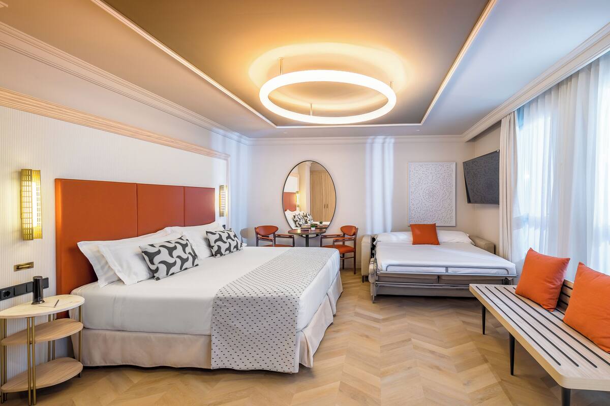 A hotel room with a large bed, a smaller bed, a desk, chairs, and a bench.