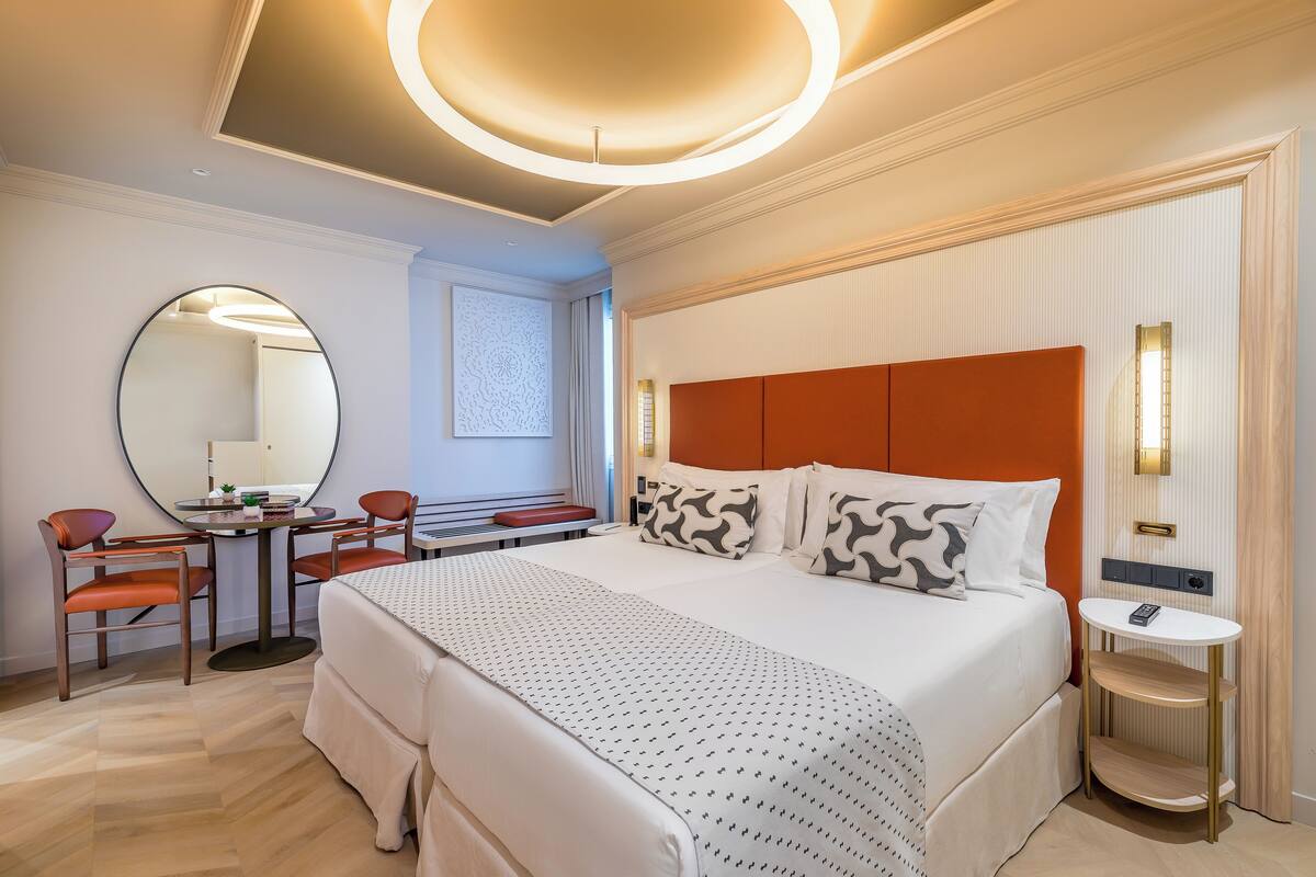 A modern hotel room with a large bed, a round mirror, a desk with chairs, and a small table with a phone.