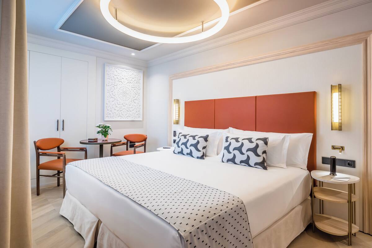 A modern hotel room with a large bed, a round ceiling light, a small table with a chair, and a window with a decorative panel.