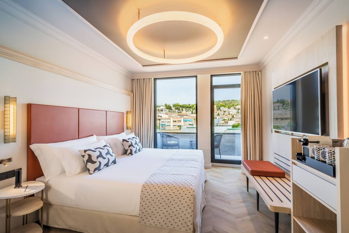 A hotel room with a large bed, a TV, a desk, and a balcony with a city view.