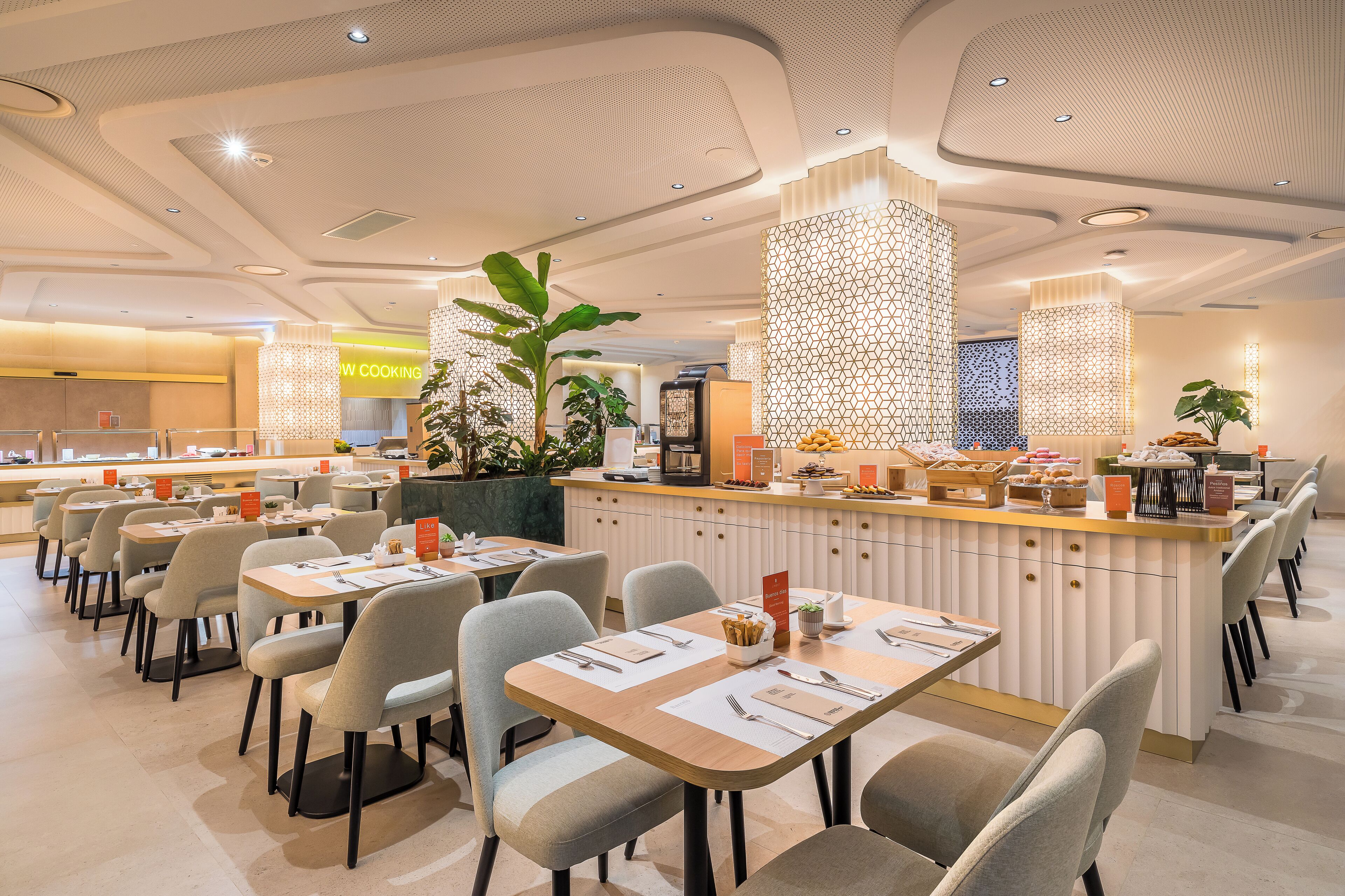 A modern dining area with tables and chairs, a central counter with food displays, and decorative plants.