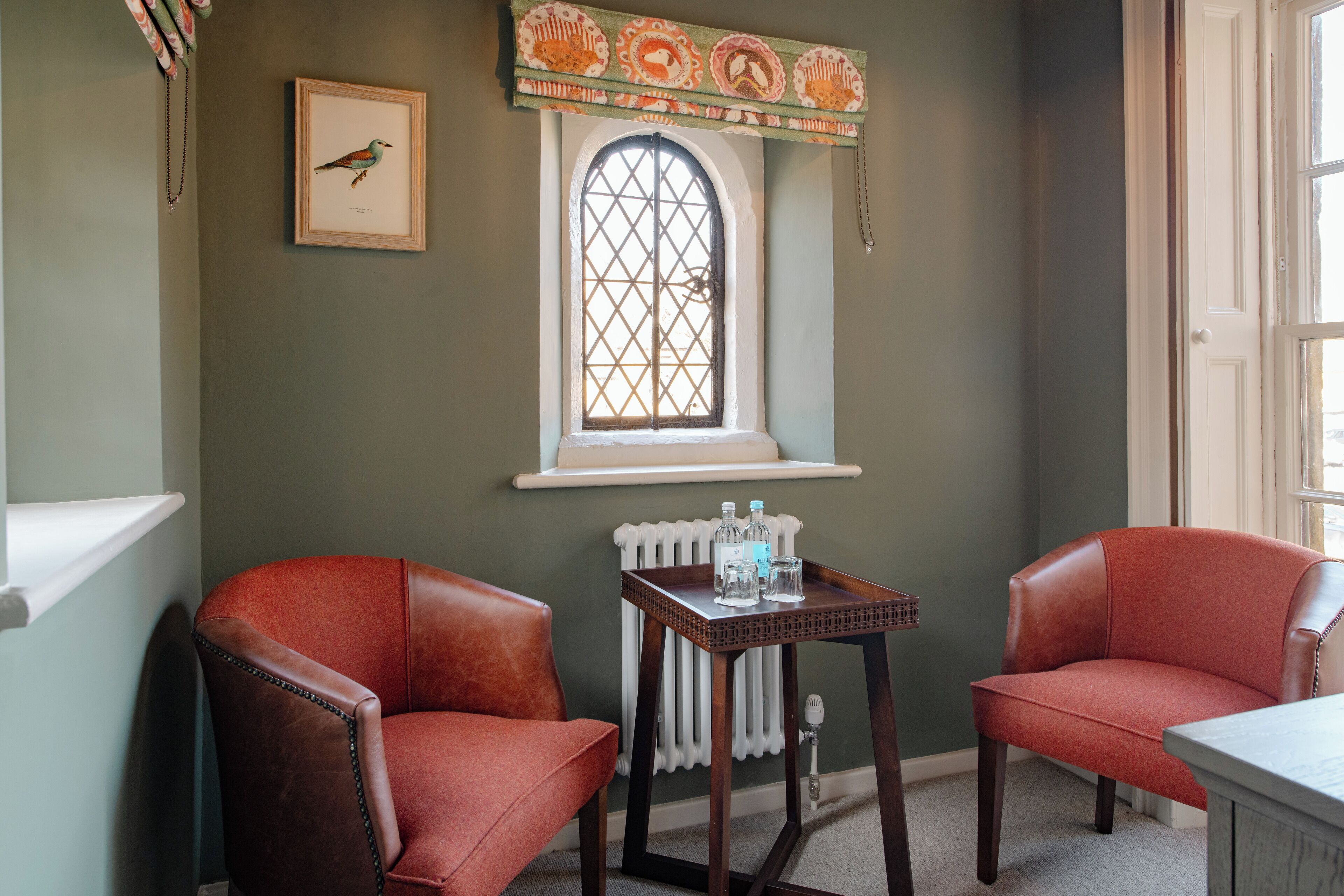 Une pièce comprenant deux fauteuils en cuir, une petite table avec des bouteilles, un tableau représentant un oiseau encadré et une fenêtre agrémentée d’une lambrequin décorative.