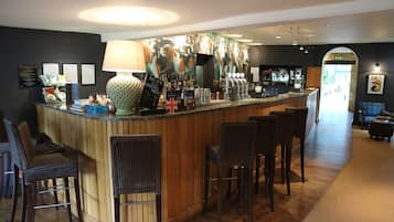 Zona de bar con mostrador de madera, taburetes y una lámpara grande. Hay diplomas enmarcados en la pared y un cartel con el menú sobre el mostrador.