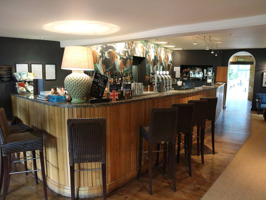 A bar area with a wooden counter, bar stools, and a large lamp. There are framed certificates on the wall and a menu sign above the counter.