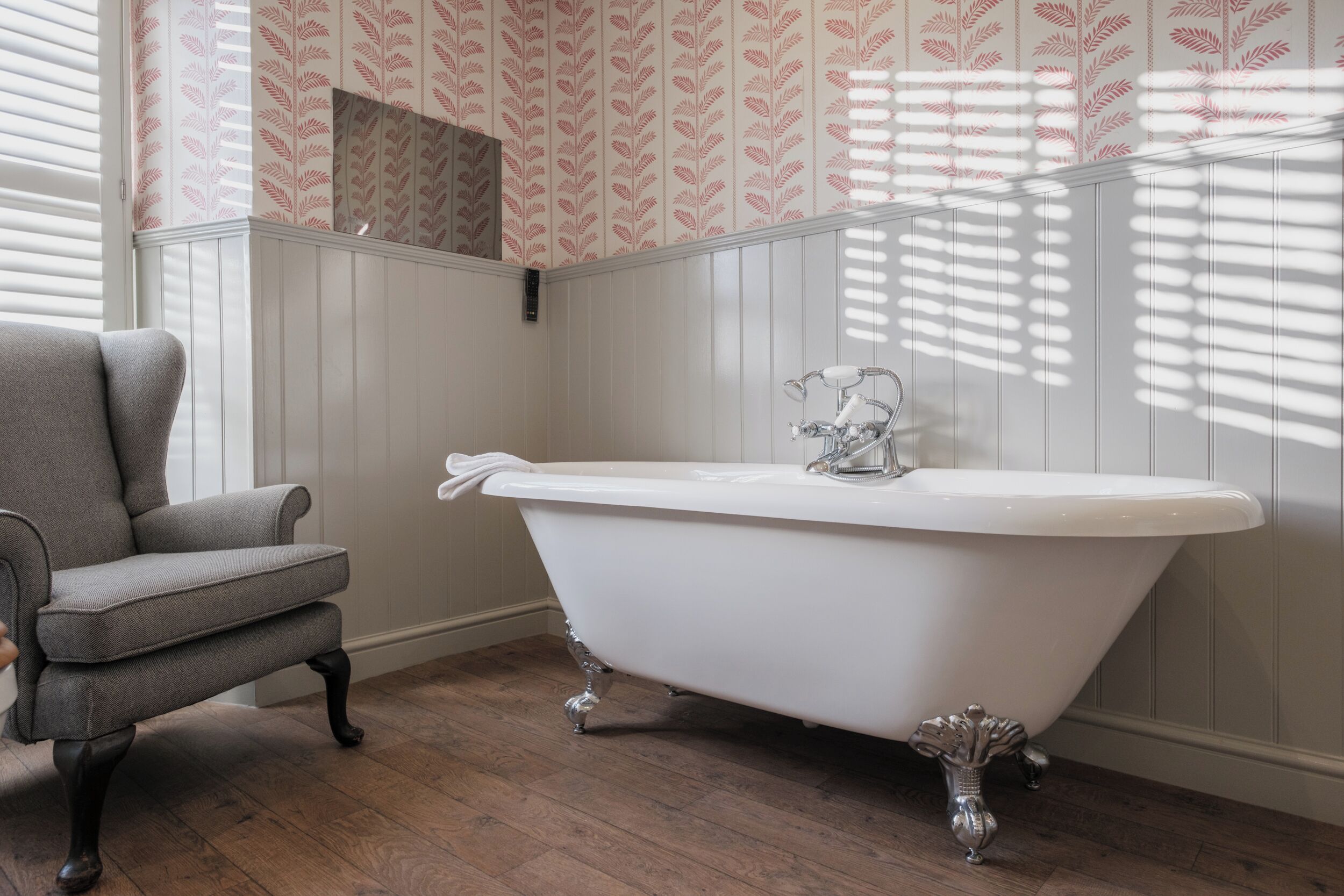 Une salle de bain avec une baignoire sur pieds, un fauteuil gris et un papier peint à motifs.