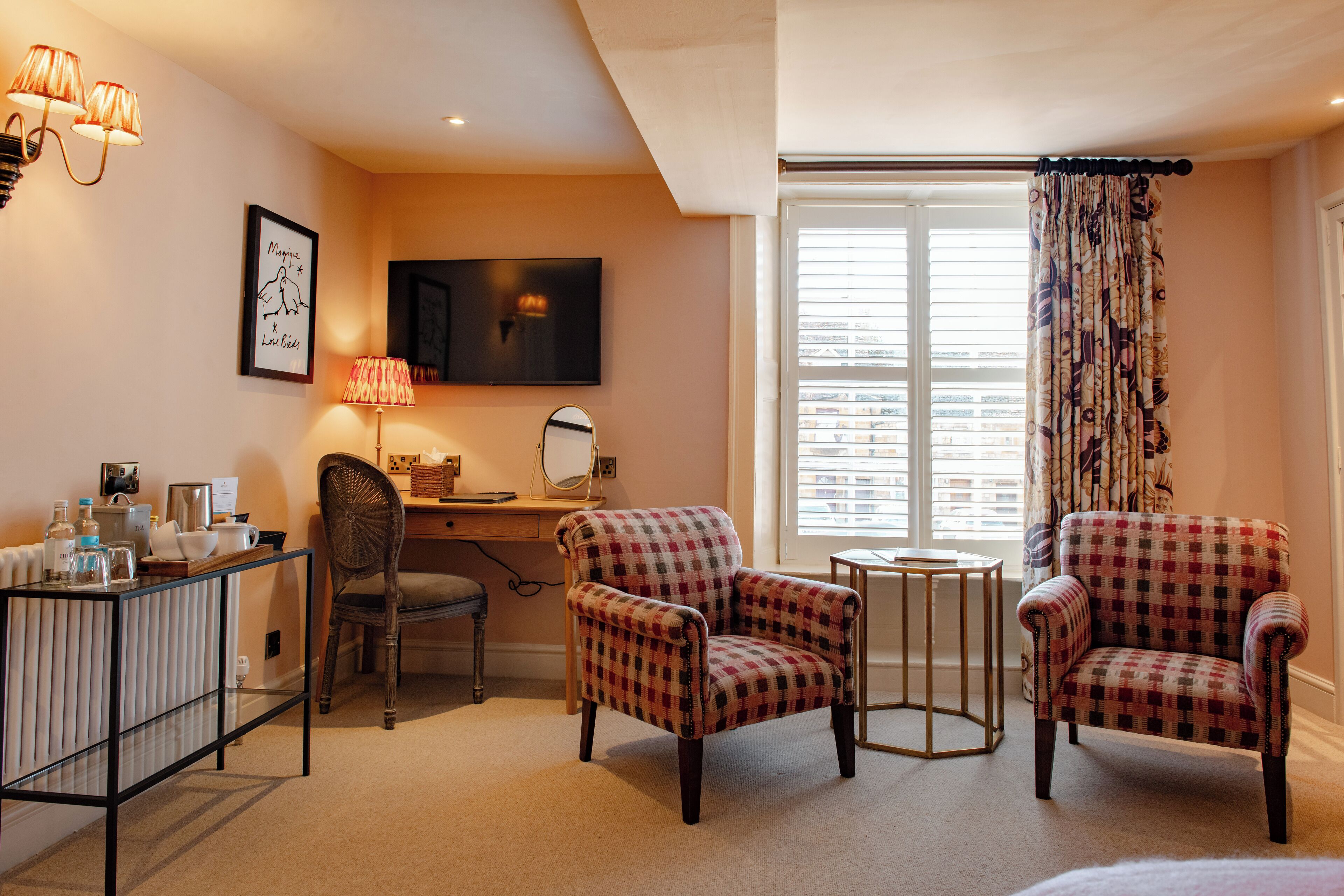 A hotel room with a desk, two armchairs, a chair, a small table, a lamp, a television, and a window with curtains.