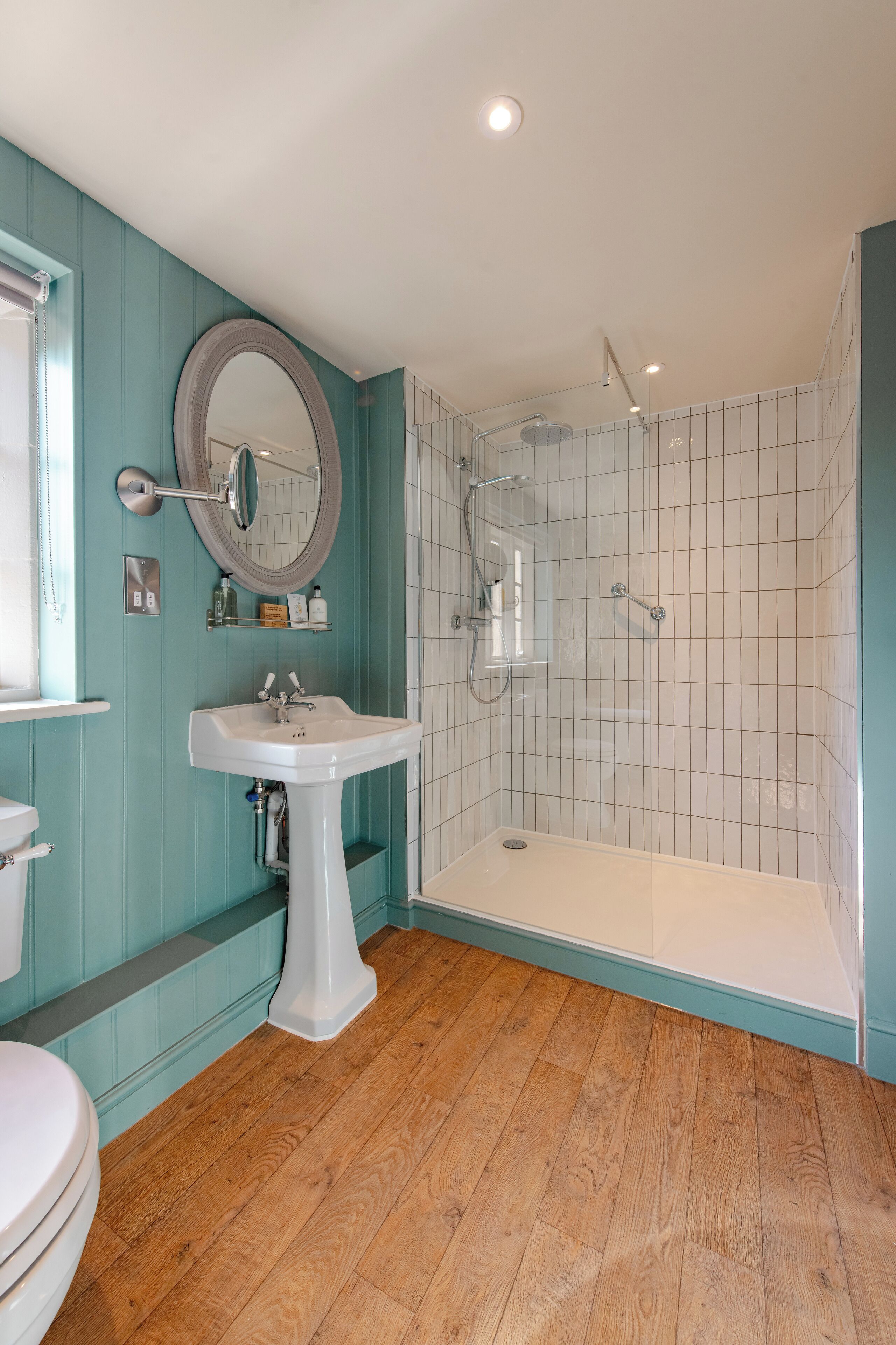 A bathroom with a white sink, a round mirror, a glass-enclosed shower, and wooden flooring.