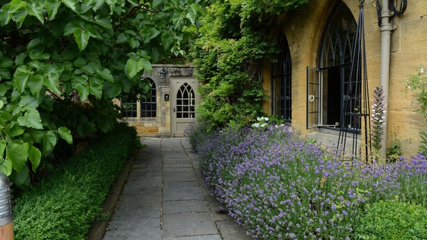 Un sentiero in pietra conduce a un edificio con finestre ad arco e un'apertura, immerso in vegetazione rigogliosa e fiori viola.