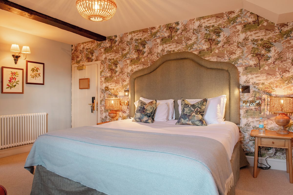 Une chambre à coucher avec un grand lit, un papier peint à motifs floraux, une tête de lit en bois, des lampes de chevet et un radiateur.