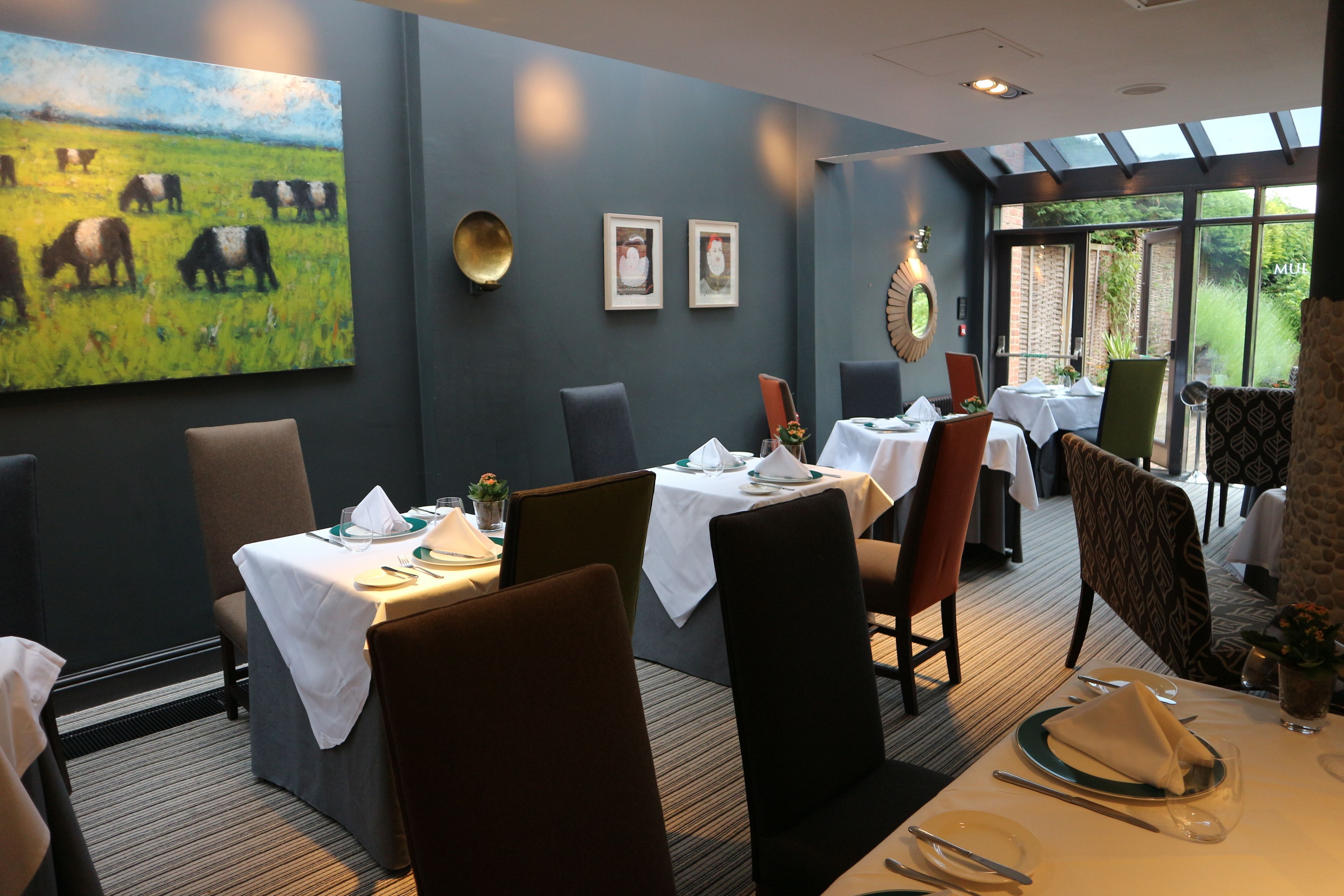 A dining area with tables set for four, chairs, and a large painting of cows on the wall.
