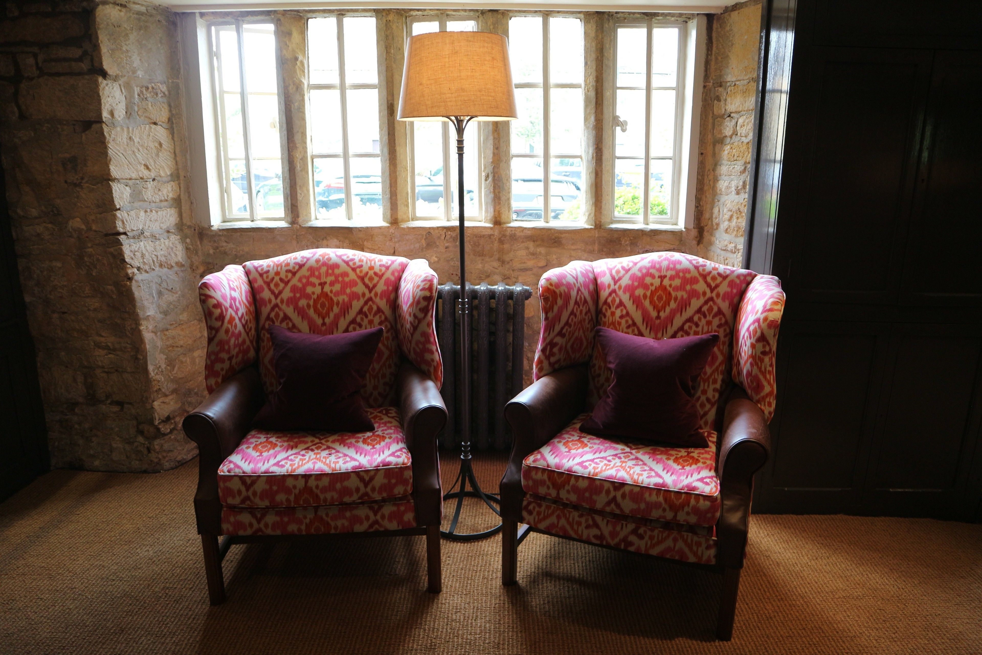 Two patterned armchairs with purple cushions in front of a radiator, near a window with a view of parked cars.