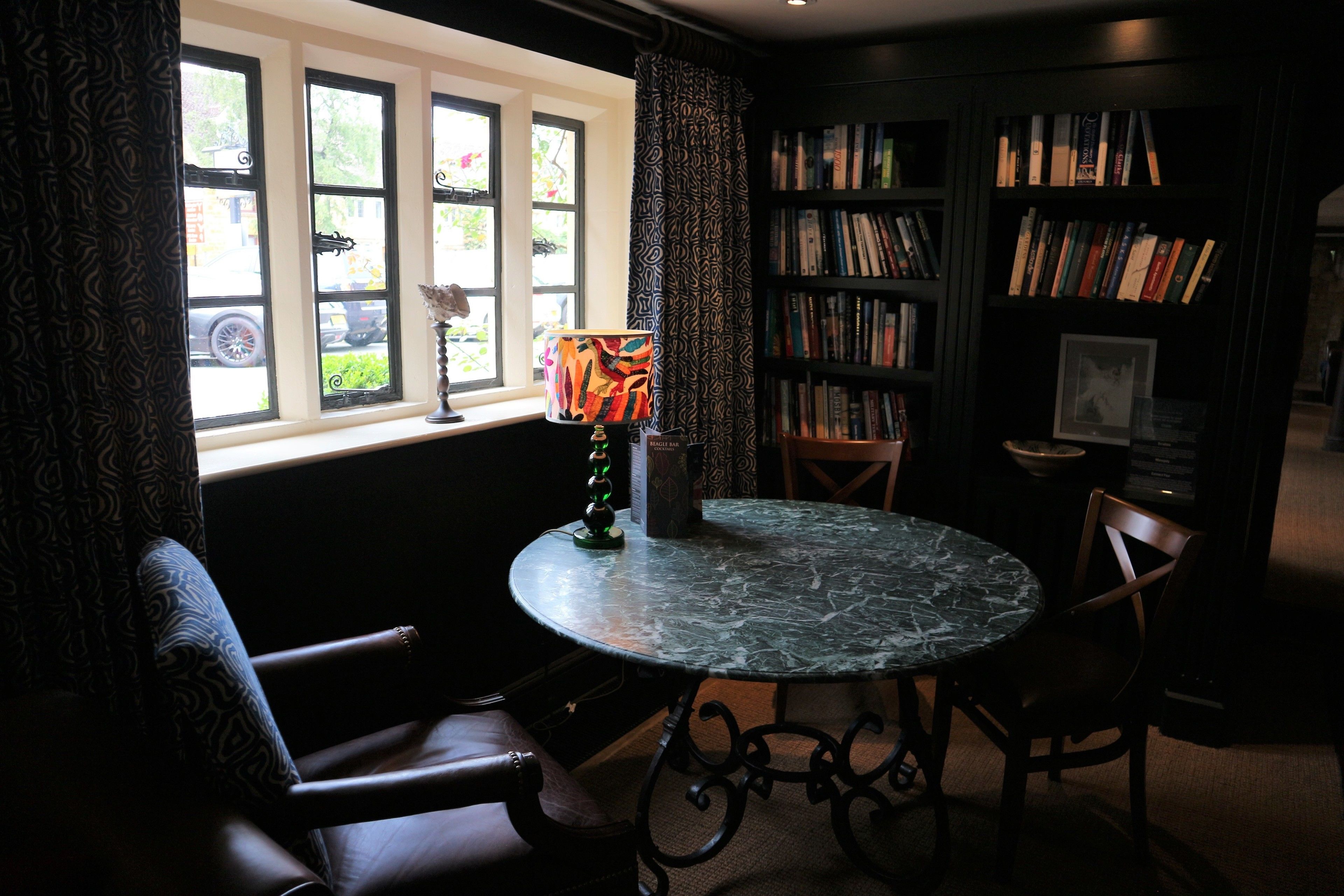 Un coin lecture confortable avec une table ronde, des fauteuils en cuir, une bibliothèque et une fenêtre avec des rideaux.
