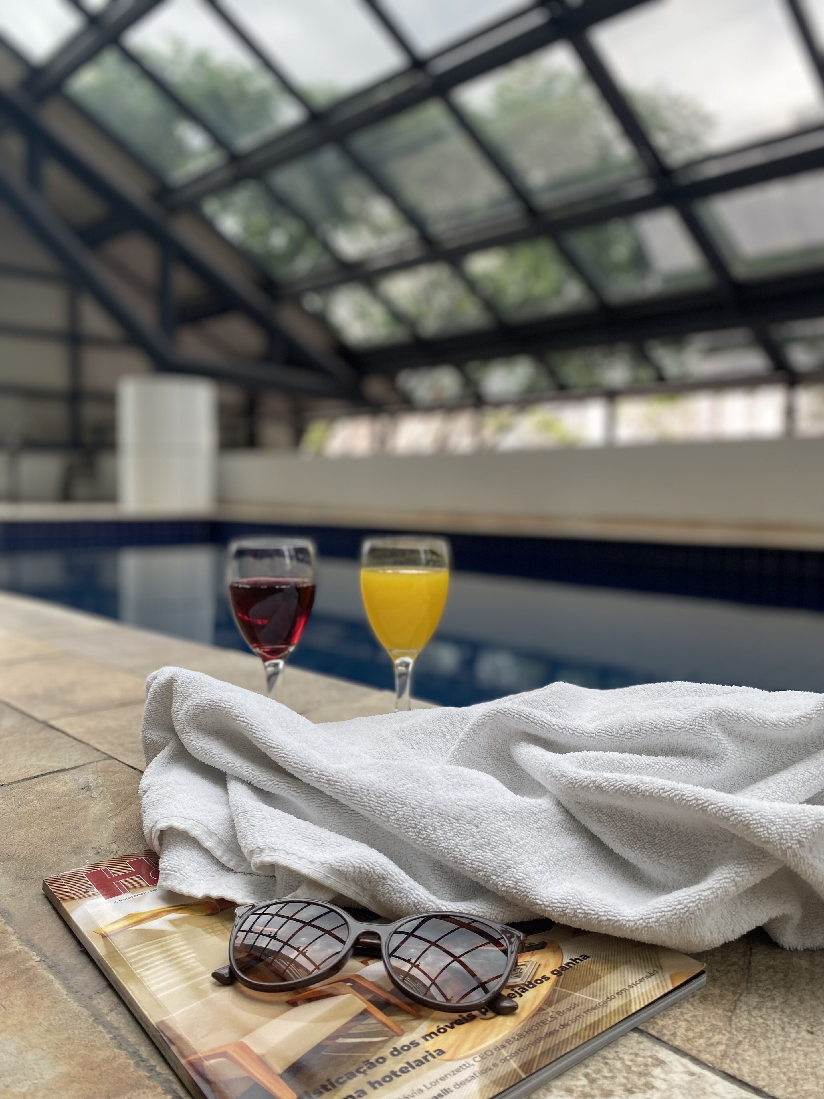 Een zwembadterras met een glas rode wijn en sinaasappelsap, een handdoek en een zonnebril op een tijdschrift.