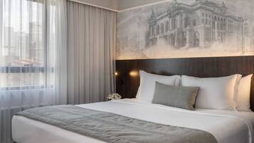 A hotel room with a large bed, a headboard with a mural, a window with sheer curtains, and a nightstand with a lamp.