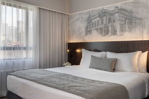 A hotel room with a large bed, a headboard with a mural, a window with sheer curtains, and a nightstand with a lamp.
