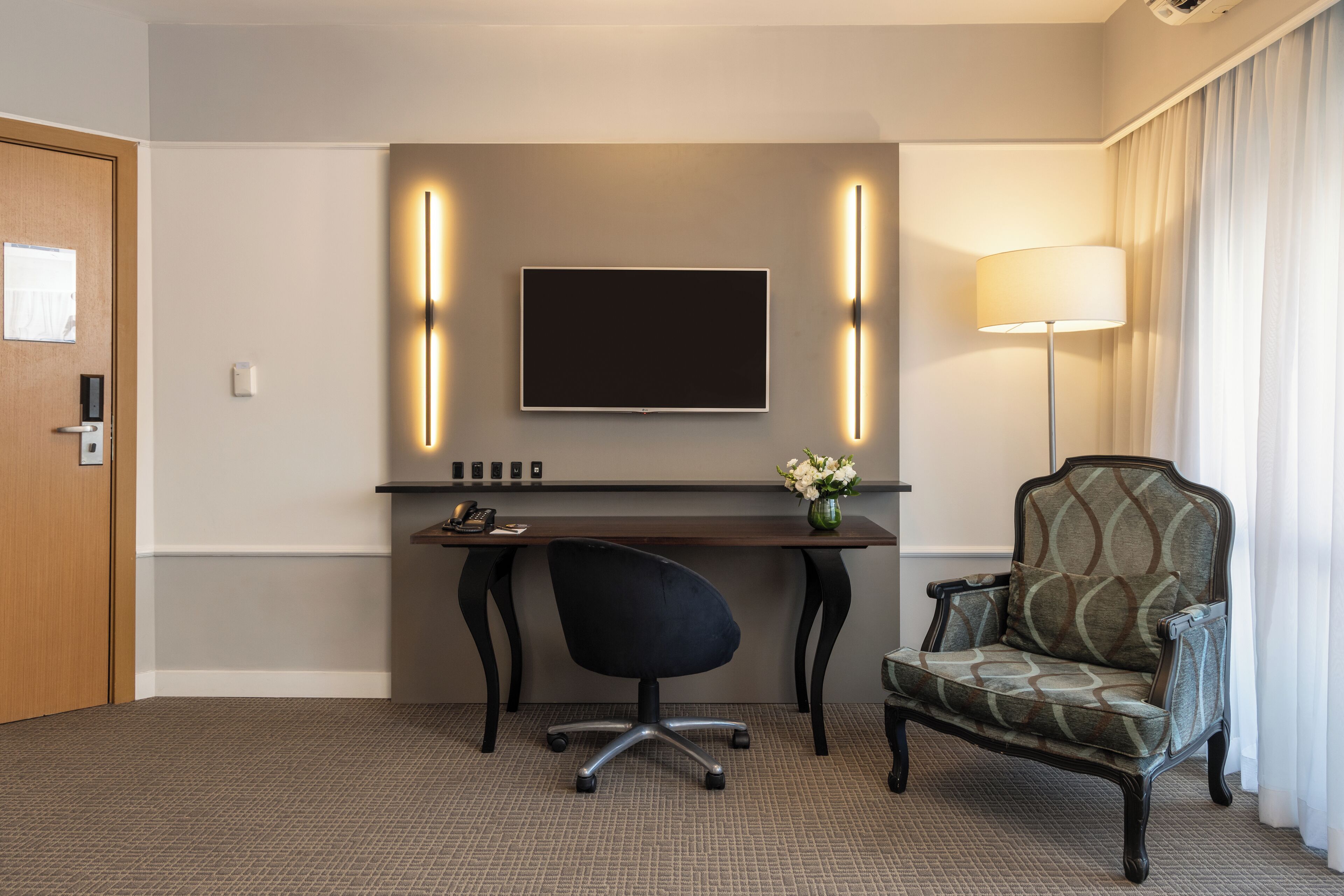 A hotel room with a desk, a chair, a flat-screen TV, and a lamp.