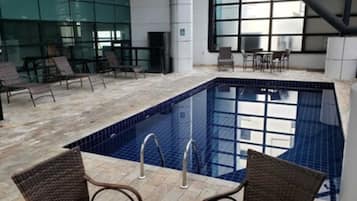 Piscina interna com paredes azulejadas e fundo azul transparente, rodeada por espreguiçadeiras e uma parede de vidro.
