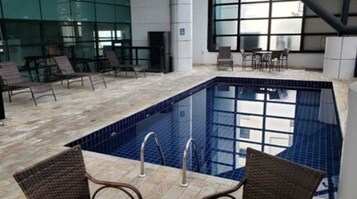 Piscina interna com paredes azulejadas e fundo azul transparente, rodeada por espreguiçadeiras e uma parede de vidro.