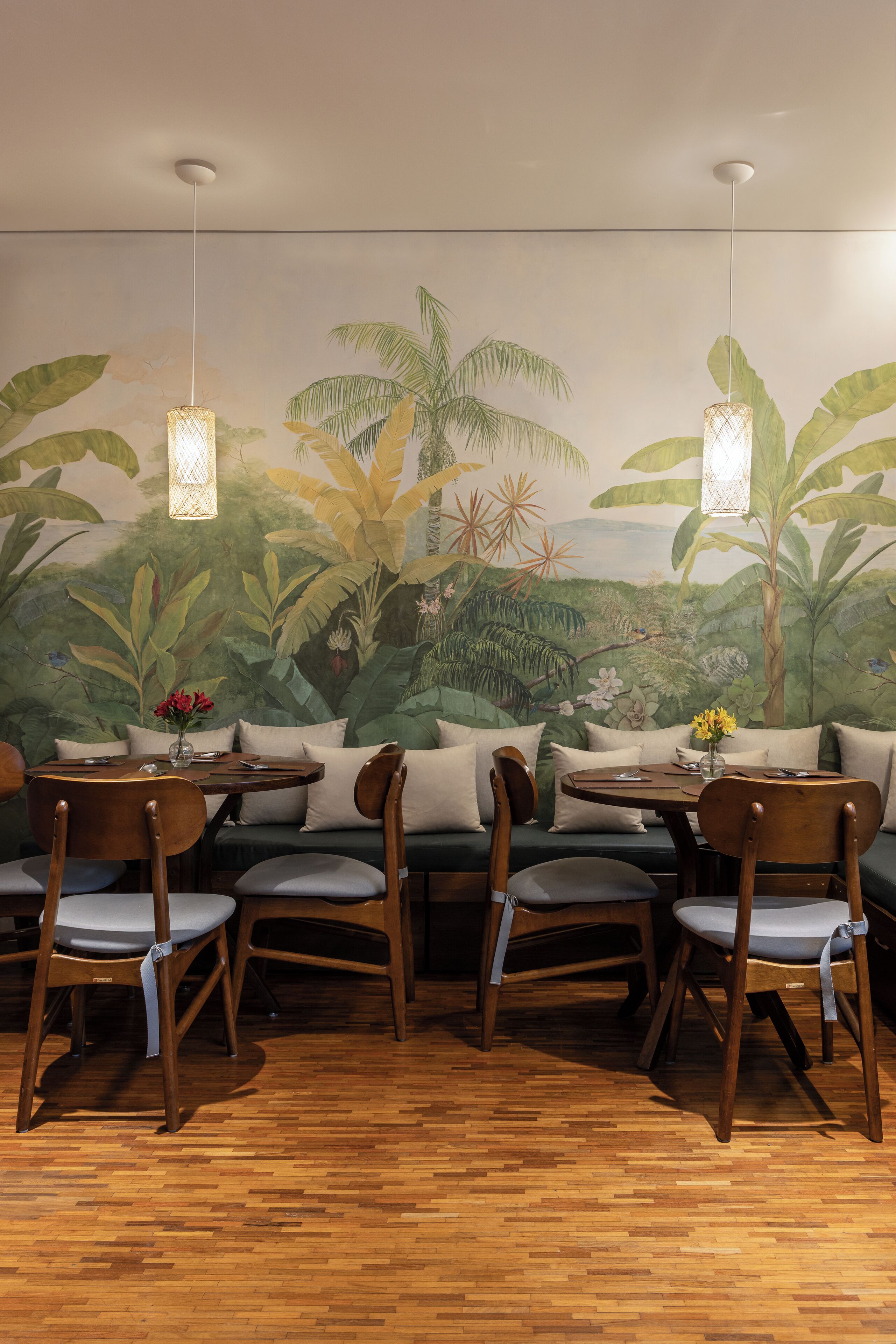 A restaurant with a mural of tropical foliage, wooden furniture, and hanging pendant lights.