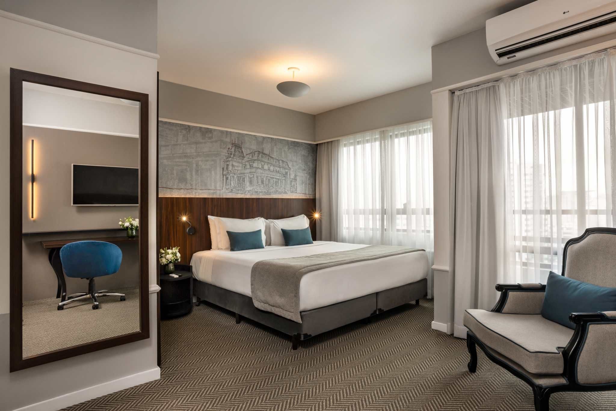 Ein Hotelzimmer mit einem großen Bett, einem Schreibtisch mit Stuhl, einem Flachbildfernseher und einem großen Fenster mit Vorhängen.