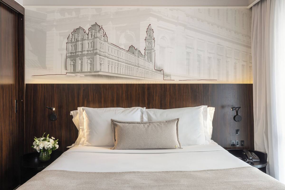 Quarto de hotel com uma cama grande, uma mesa de cabeceira com abajur, telefone e uma obra de arte na parede retratando um edifício.