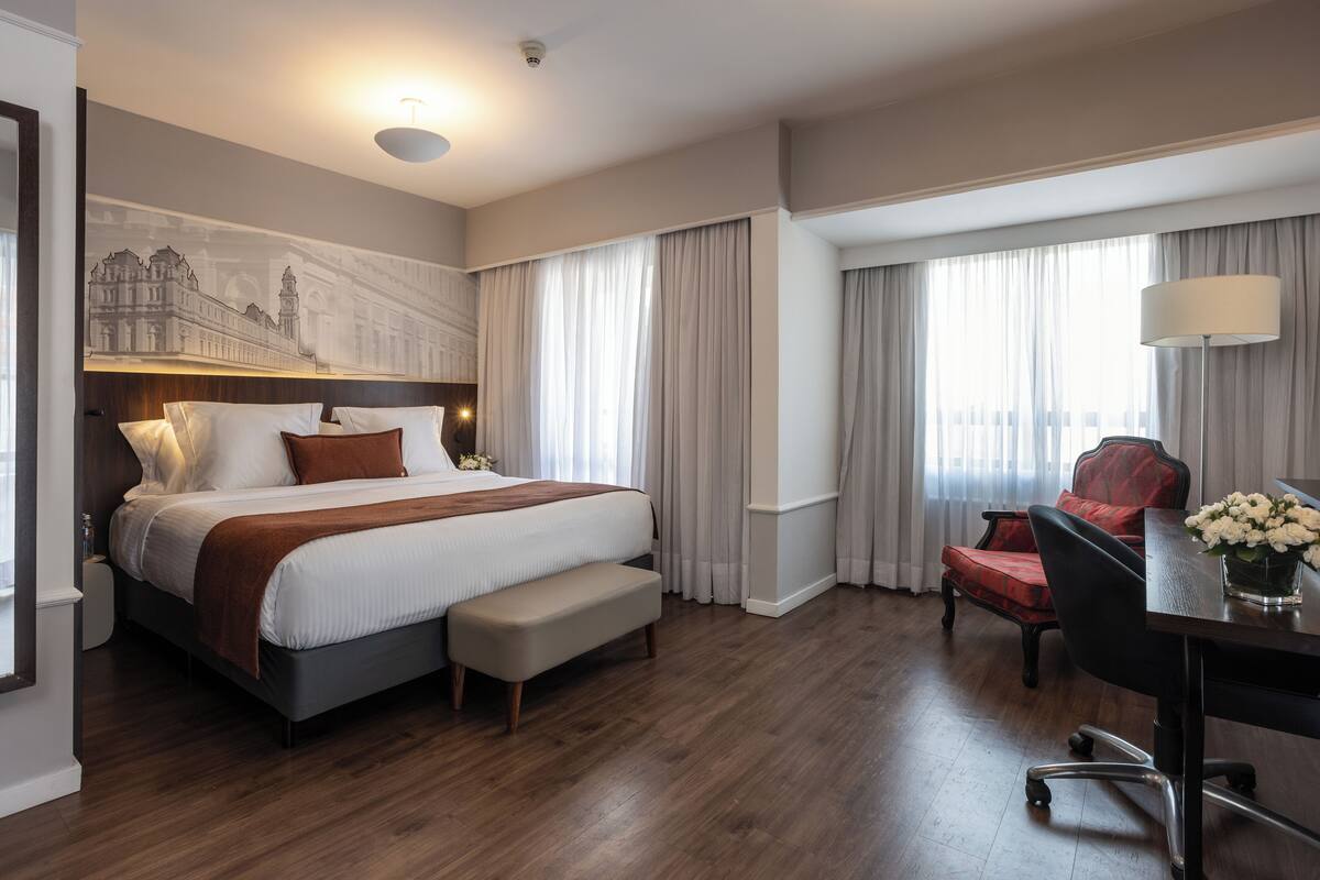 Quarto de hotel com cama grande, escrivaninha com cadeira, um banco e uma janela ampla com cortinas.