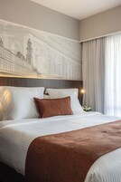A hotel room with a large bed, white bedding, a brown throw blanket, and a wall-mounted artwork of a building.